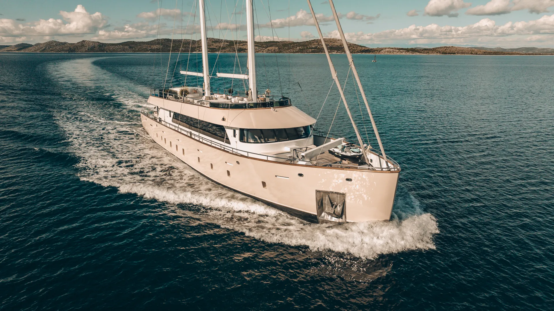 MAXITA A large white luxury yacht sails through calm blue waters, leaving a gentle wake behind. Hills and a partly cloudy sky are visible in the background.