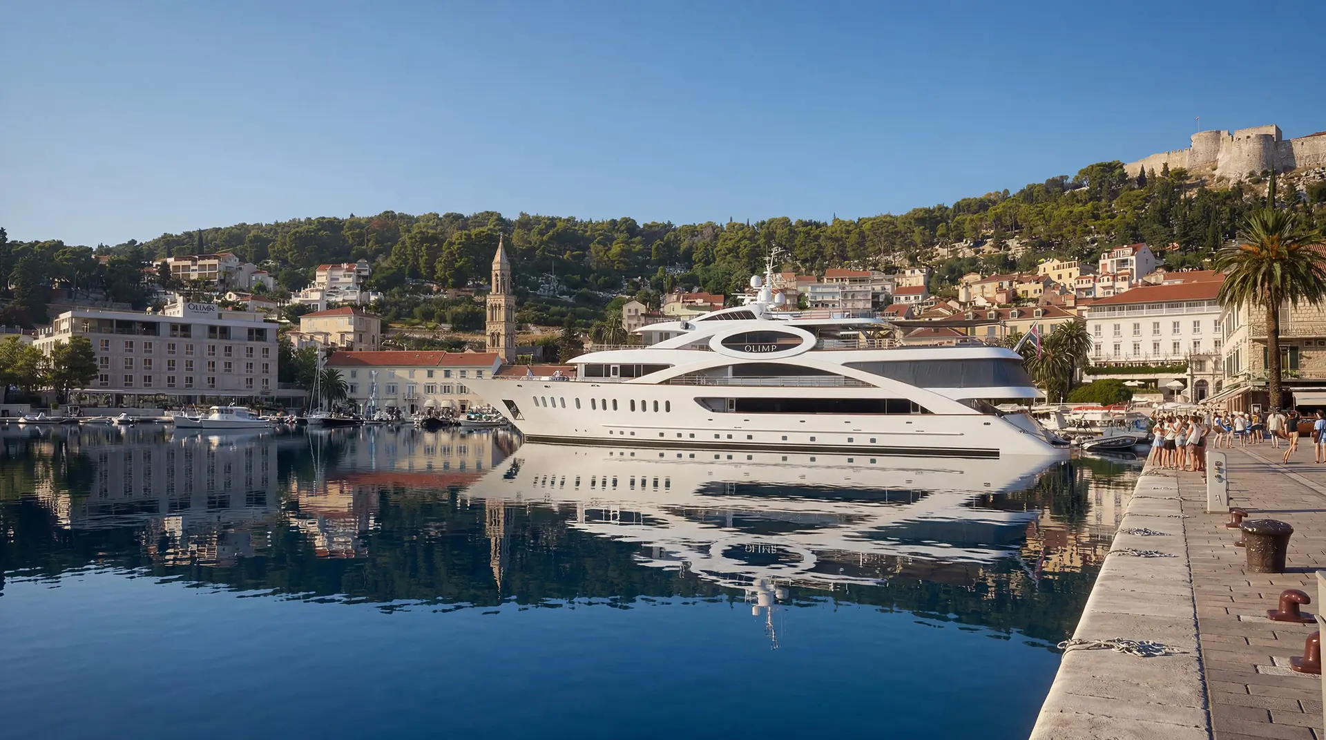 A large white yacht is docked in a calm harbor, reflecting in the water. Colorful buildings, trees, and hills are in the background under a clear blue sky, with a stone promenade on the right.