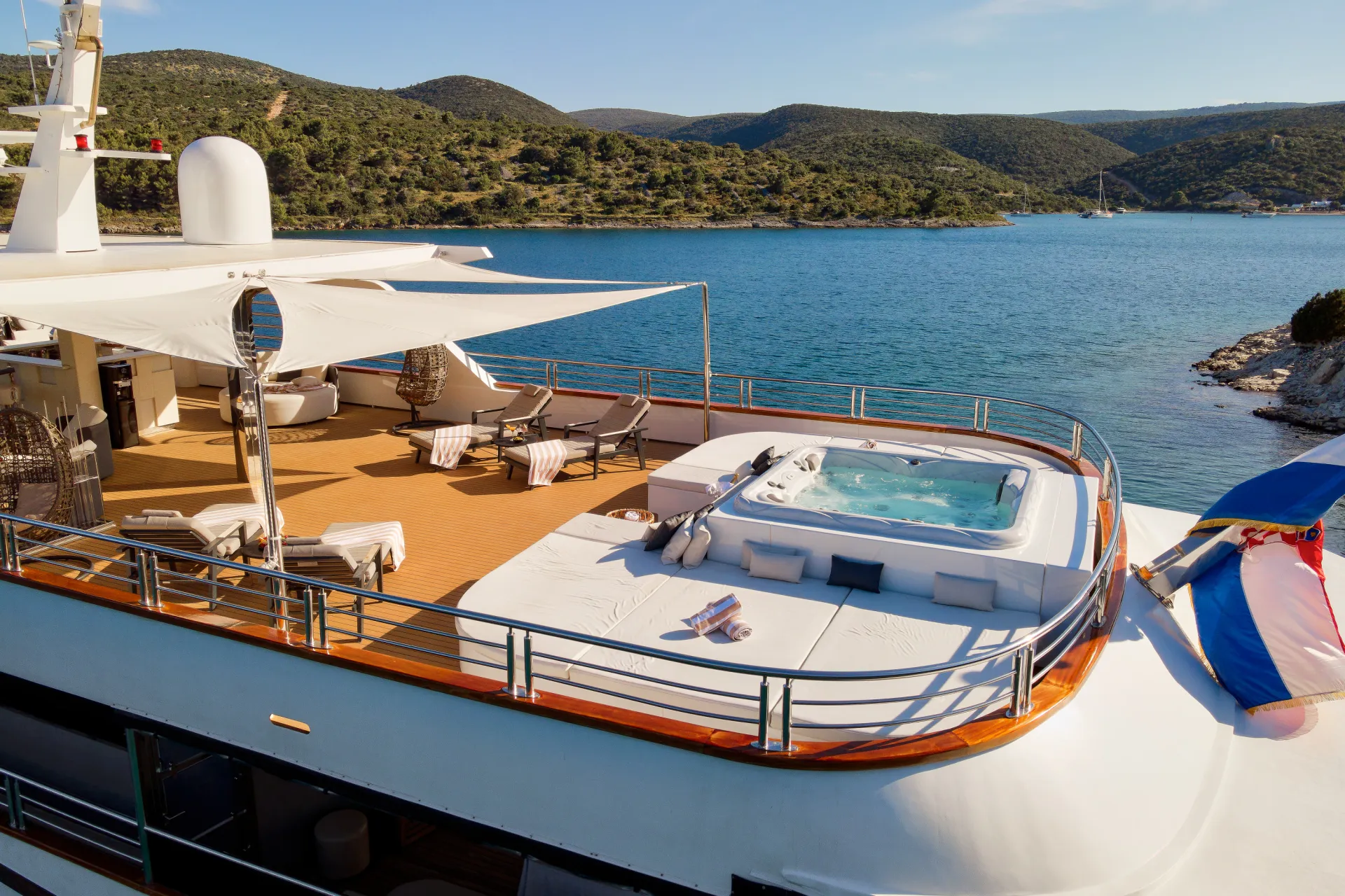 CRISTAL A luxury yacht deck features sun loungers, hanging chairs, and a large jacuzzi overlooking calm blue water and green hills under a clear sky. Towels and cushions are neatly arranged for relaxation.