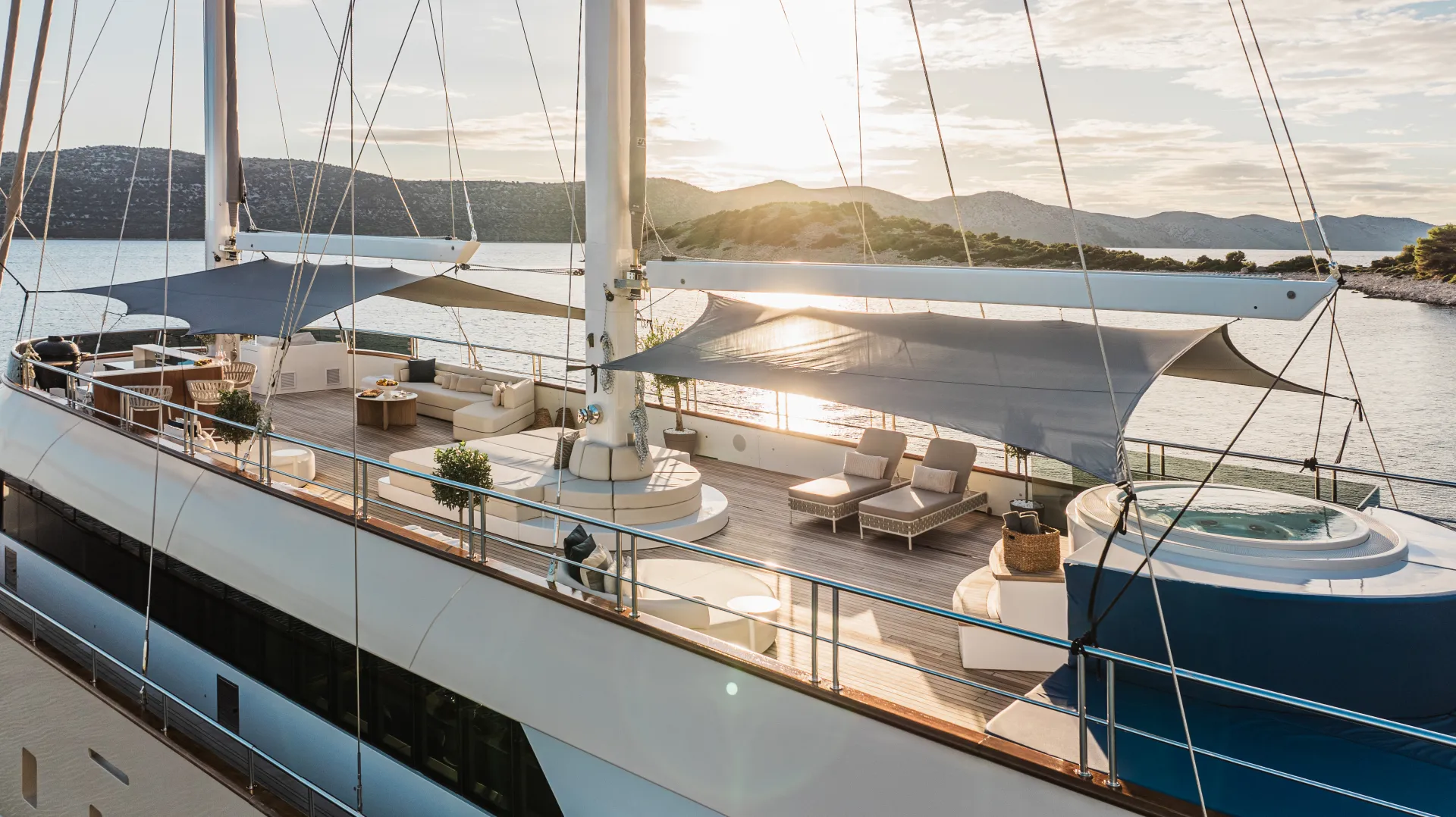MAXITA A luxurious yacht deck with lounge chairs, sofas, shaded areas, potted plants, and a jacuzzi overlooks calm water and distant hills at sunset.