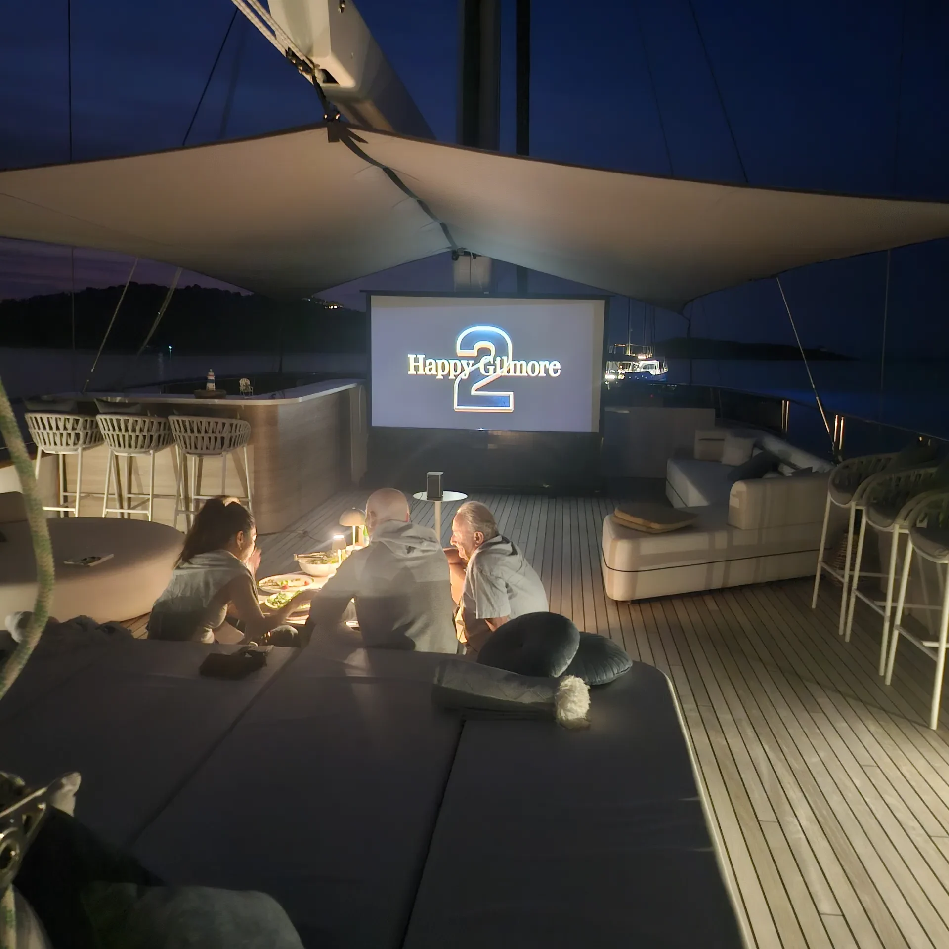 MAXITA A group of people sits on a boat deck at night, watching a movie projected on a screen that reads Happy 2nd move. Cozy seating and dim lighting create a relaxed atmosphere.