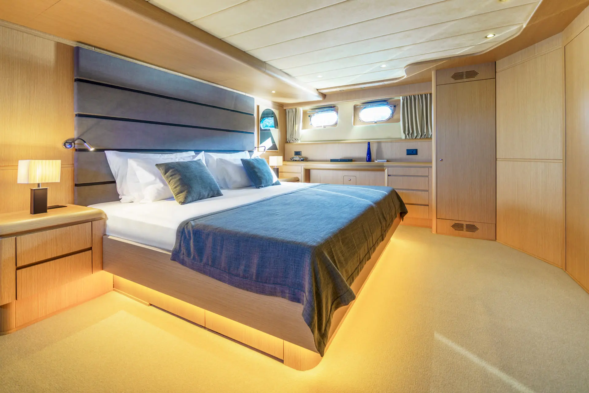 Modern yacht bedroom with a large bed, blue bedding, two bedside tables with lamps, wood-paneled walls, built-in storage, and small windows letting in natural light. Warm lighting is under the bed and along the ceiling.