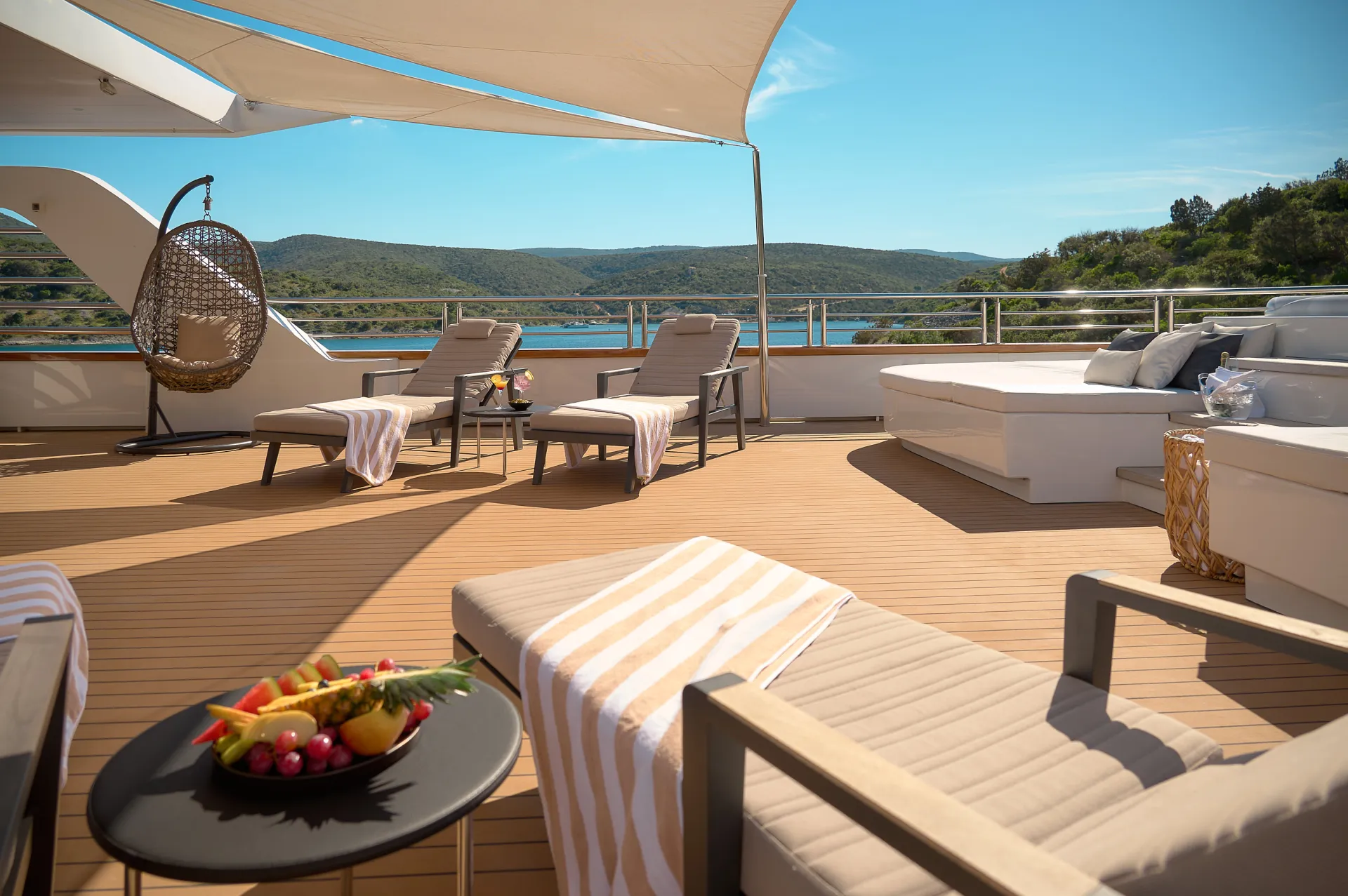 CRISTAL A luxurious yacht deck with lounge chairs, a round daybed, a hanging chair, and a table with fresh fruit. The scene overlooks calm blue water and green hills under a sunny sky.