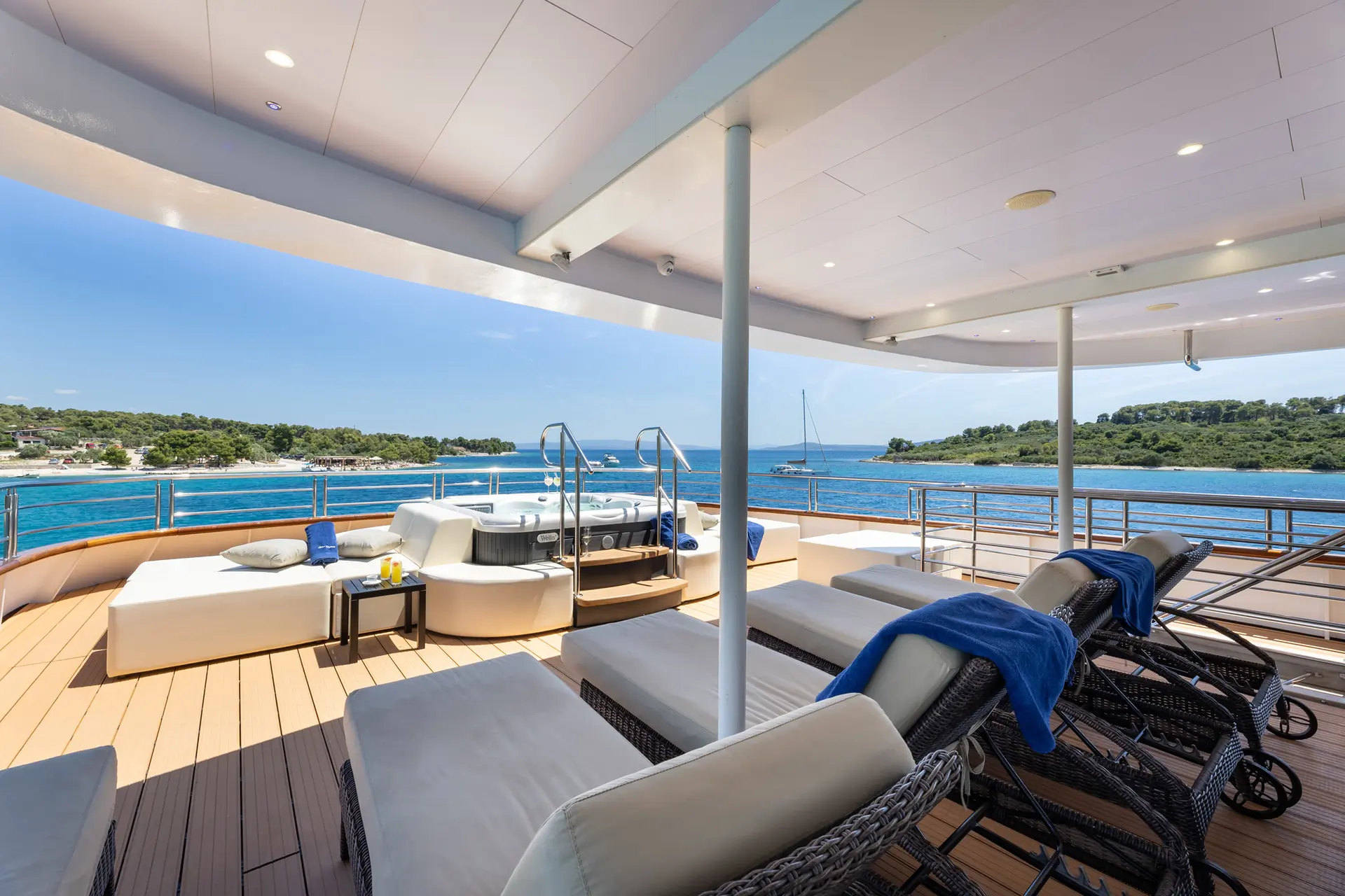 Lounge chairs and a hot tub on the sunny deck of a luxury yacht overlook a blue ocean and distant green shoreline under a clear sky.