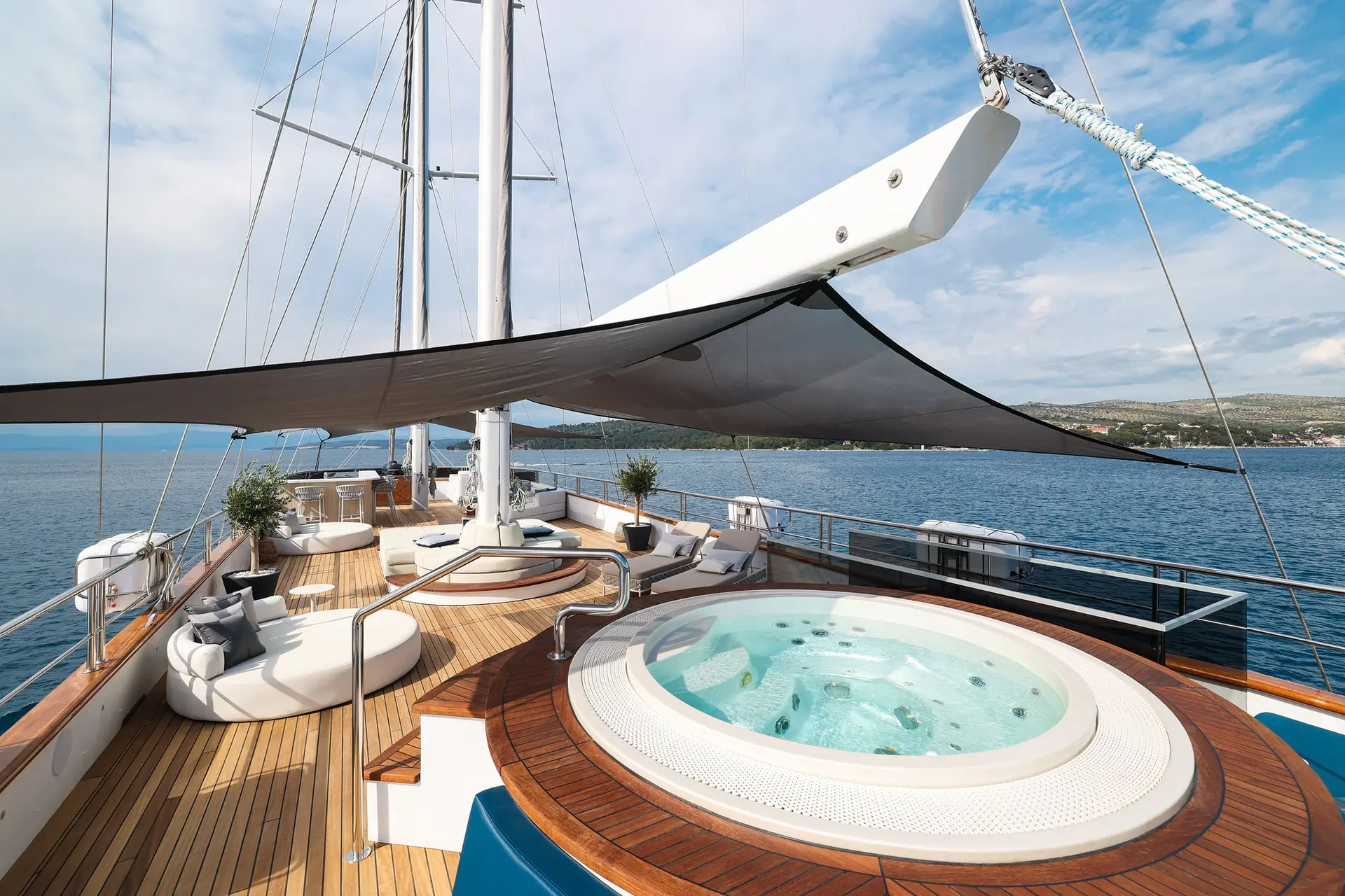 A luxury yacht deck with a round hot tub, modern seating, potted plants, and a large sunshade, overlooking calm blue water and a distant shore under a partly cloudy sky.