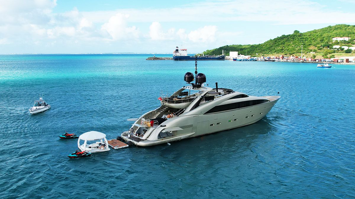 YCM 120 A large luxury yacht is anchored in clear blue water near a green coastline, with smaller boats and jet skis floating nearby. The sky is partly cloudy, and there are buildings and a harbor in the distance.
