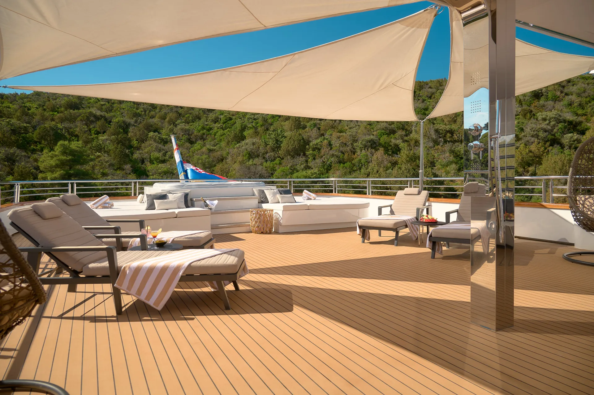 CRISTAL A spacious yacht deck with lounge chairs, cushioned seating, tables with drinks, and a sunshade. Lush green hills and a clear blue sky are visible in the background.