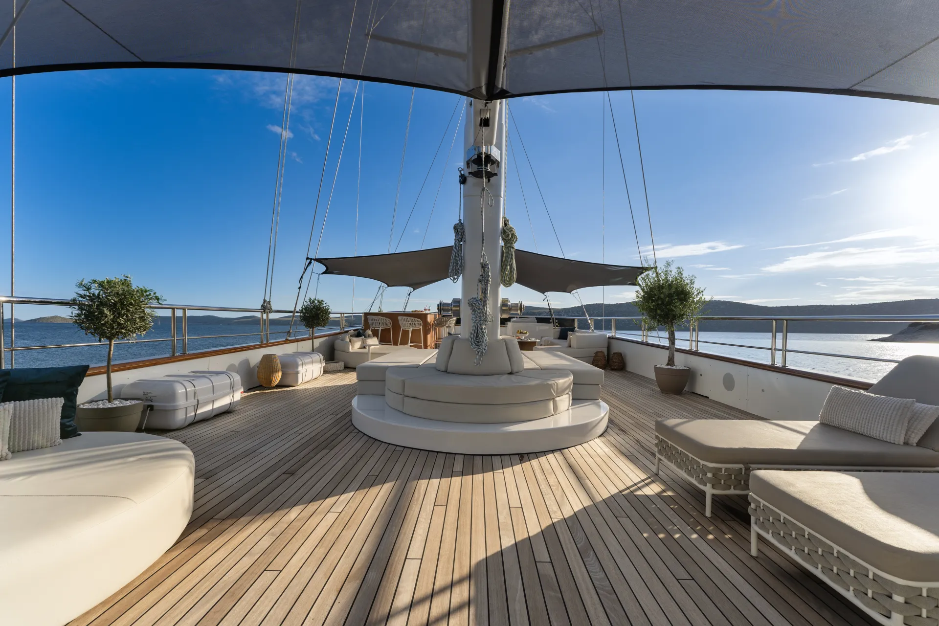 MAXITA Spacious yacht deck with wooden flooring, white cushioned seating, sun loungers, potted plants, and shade sails, overlooking calm blue water and distant hills under a clear sky.