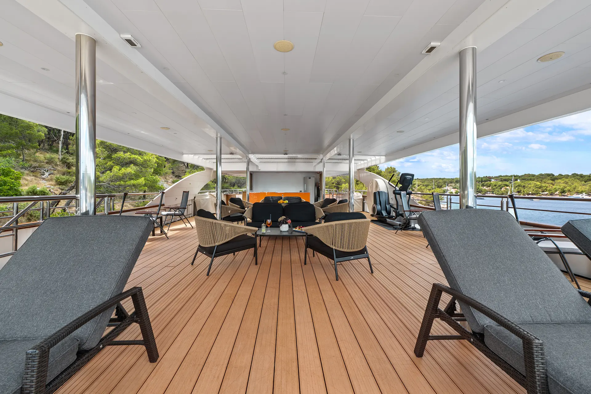 Spacious yacht deck with lounge chairs, a seating area with a table, and exercise equipment, overlooking water and trees on a sunny day.