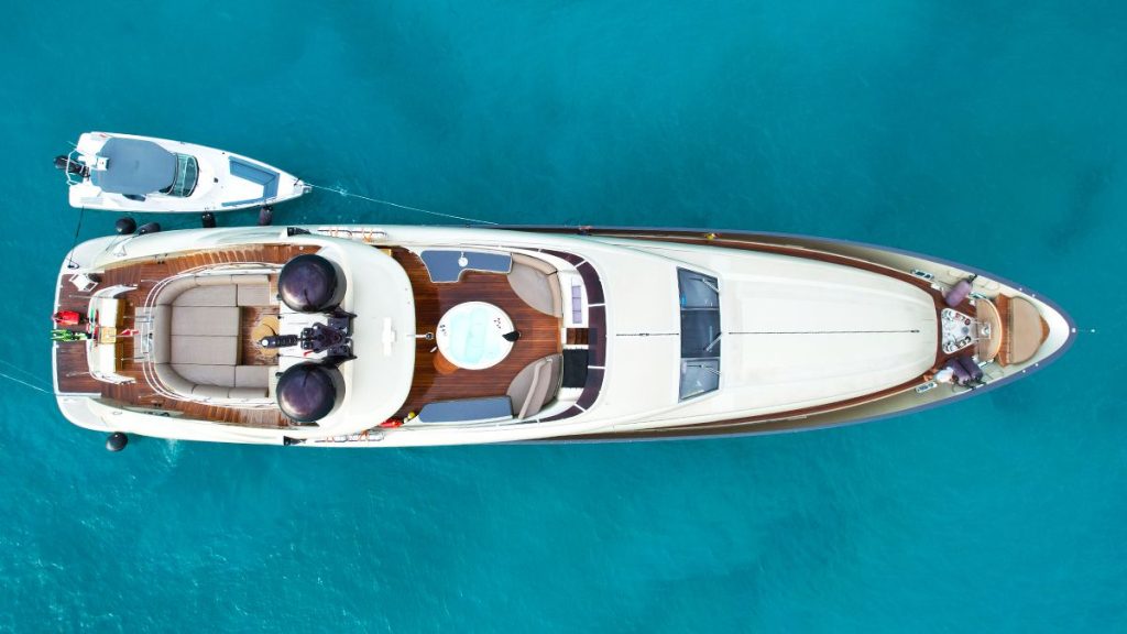 YCM 120 Aerial view of a luxury yacht with a small boat tethered to it, floating on clear turquoise water. The yacht has sun loungers and seating areas on its deck.
