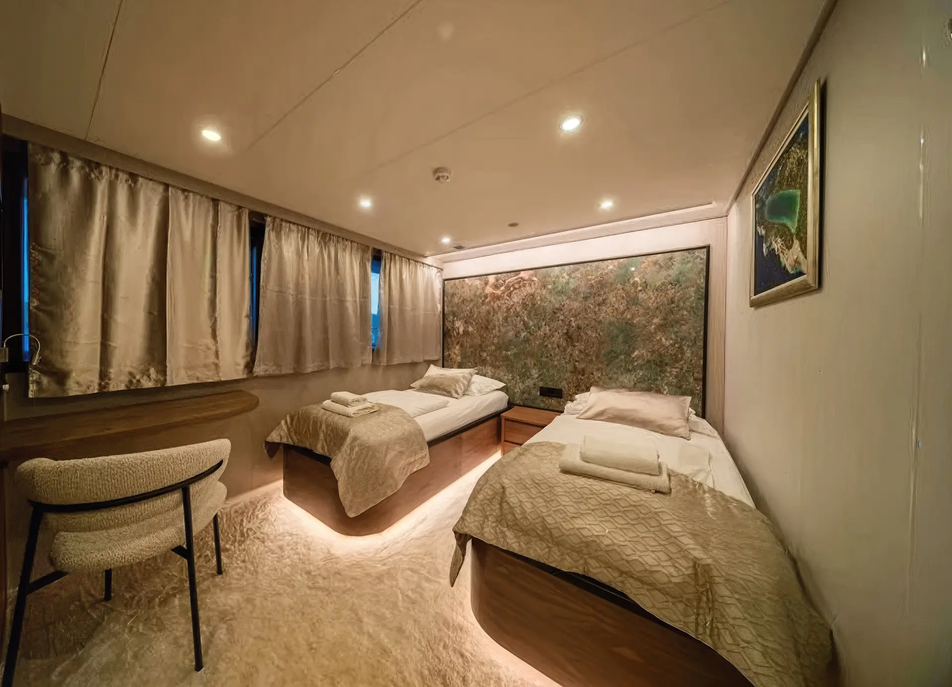 A cozy modern bedroom with two single beds, beige bedding, folded towels, a soft rug, a desk with a chair, curtained windows, a textured accent wall, and a framed map on the wall.