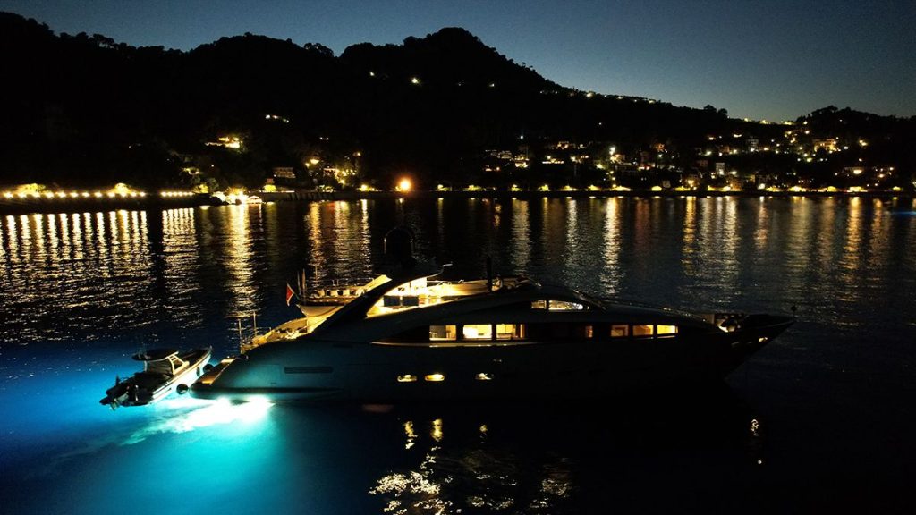 YCM 120 A luxury yacht with underwater lights is anchored near a coastline at night, with hills and shoreline buildings illuminated in the background and their reflections shimmering on the water.
