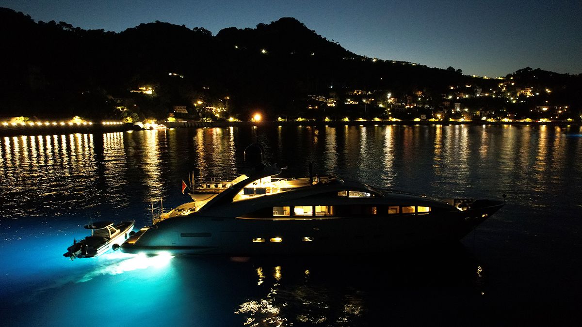 YCM 120 A luxury yacht with underwater lights is anchored near a coastline at night, with hills and shoreline buildings illuminated in the background and their reflections shimmering on the water.