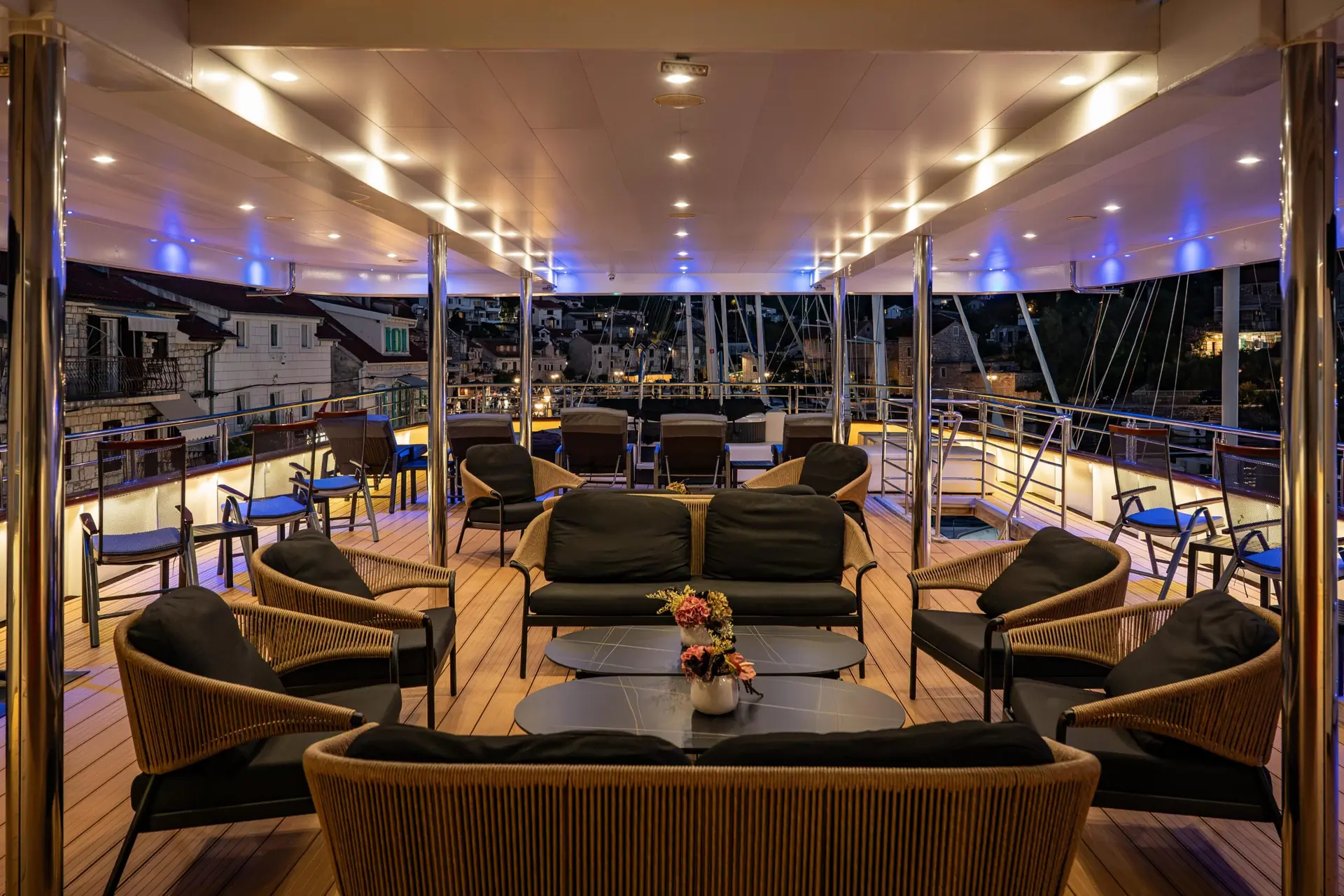 Outdoor deck of a yacht at night with cushioned seating, tables, and soft lighting. There are flowers on the tables, and buildings with lit windows are visible in the background. The scene is elegant and inviting.
