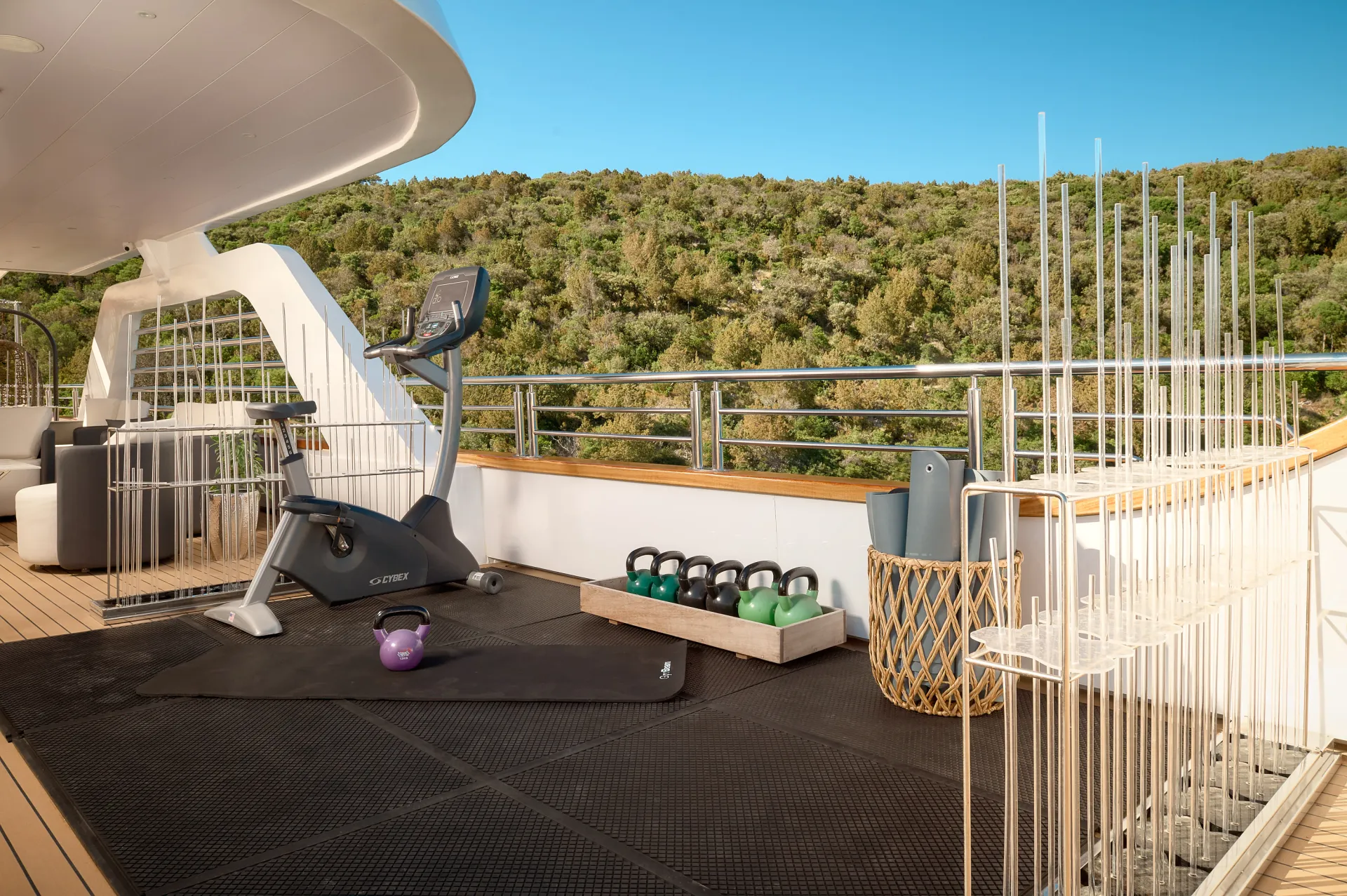 CRISTAL A yacht deck gym with an exercise bike, kettlebells, yoga mats, and scenic greenery in the background under a clear blue sky.