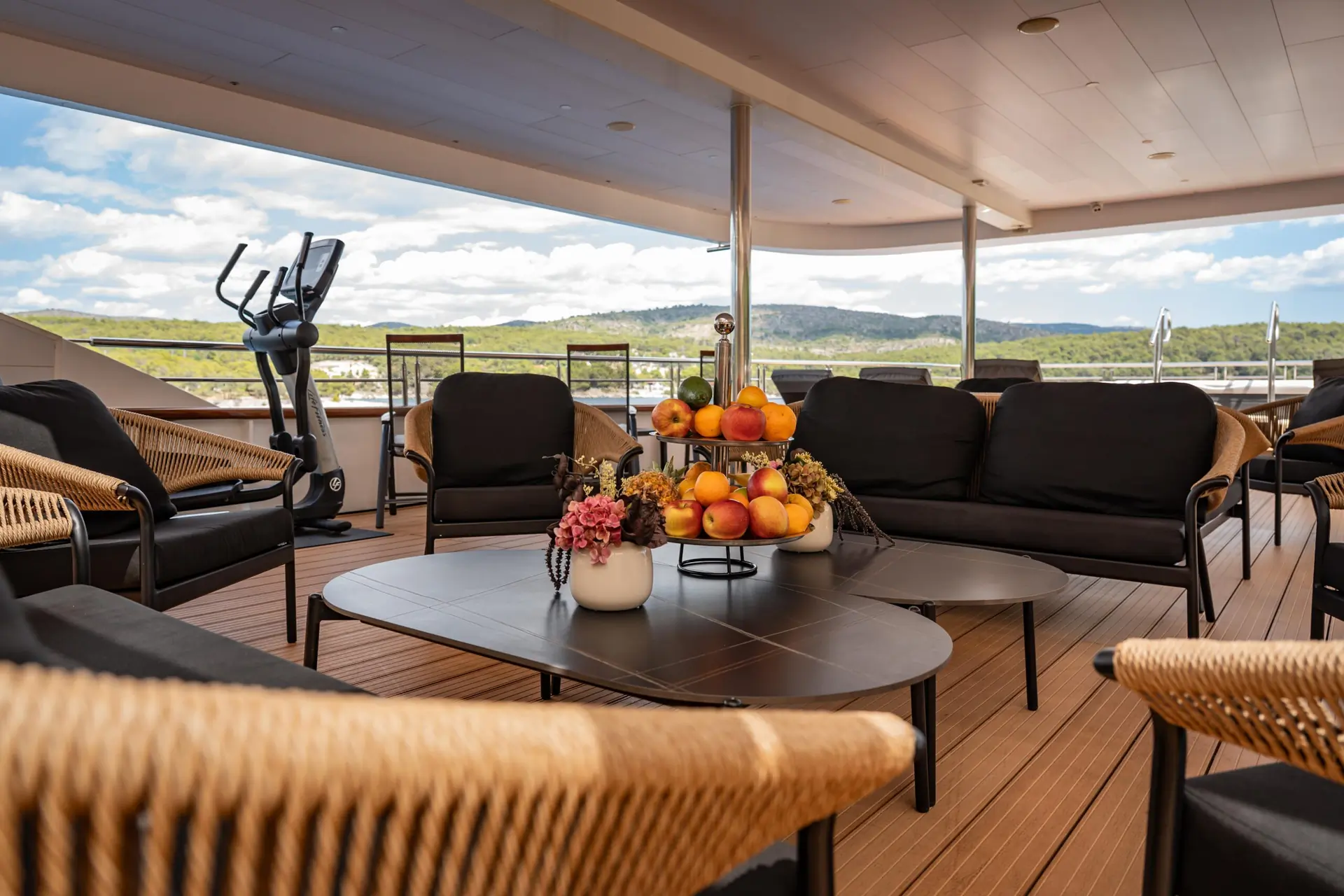 A spacious yacht deck with black cushioned seating, a table with a fruit centerpiece, potted flowers, an exercise bike, and scenic views of hills and clouds in the background.