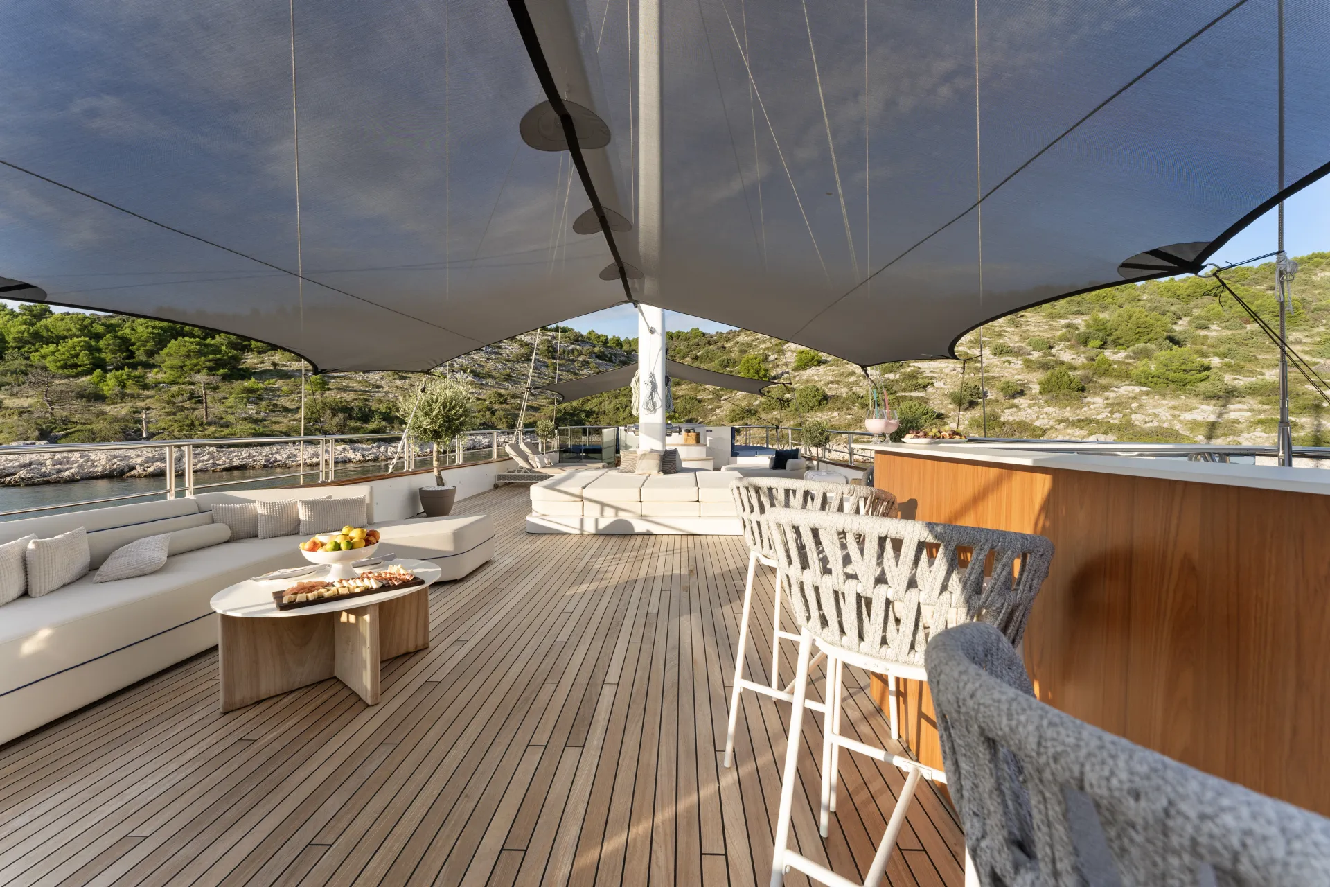 MAXITA Luxurious yacht deck with modern seating, tables with food, wooden flooring, and a sleek bar area under a large sunshade, set against a scenic coastal landscape with greenery and cliffs.
