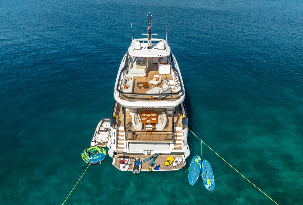 VIVA II Aerial view of a luxury yacht anchored in clear blue water, with water toys such as kayaks, paddleboards, and a small boat arranged at the stern. The yacht has multiple decks with outdoor seating and sunbathing areas.