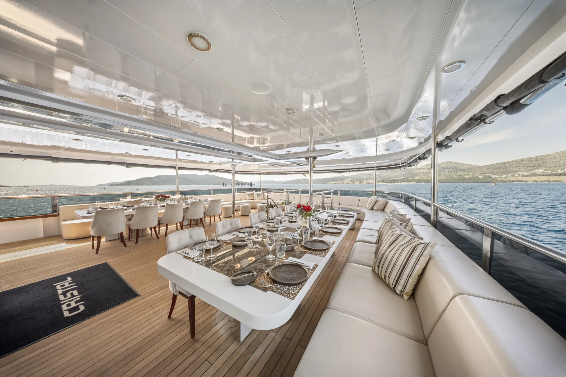 CRISTAL A luxury yacht deck with two elegant dining areas set for meals, featuring white tables, cushioned chairs, and a built-in cream sofa; ocean and distant hills visible in the background.