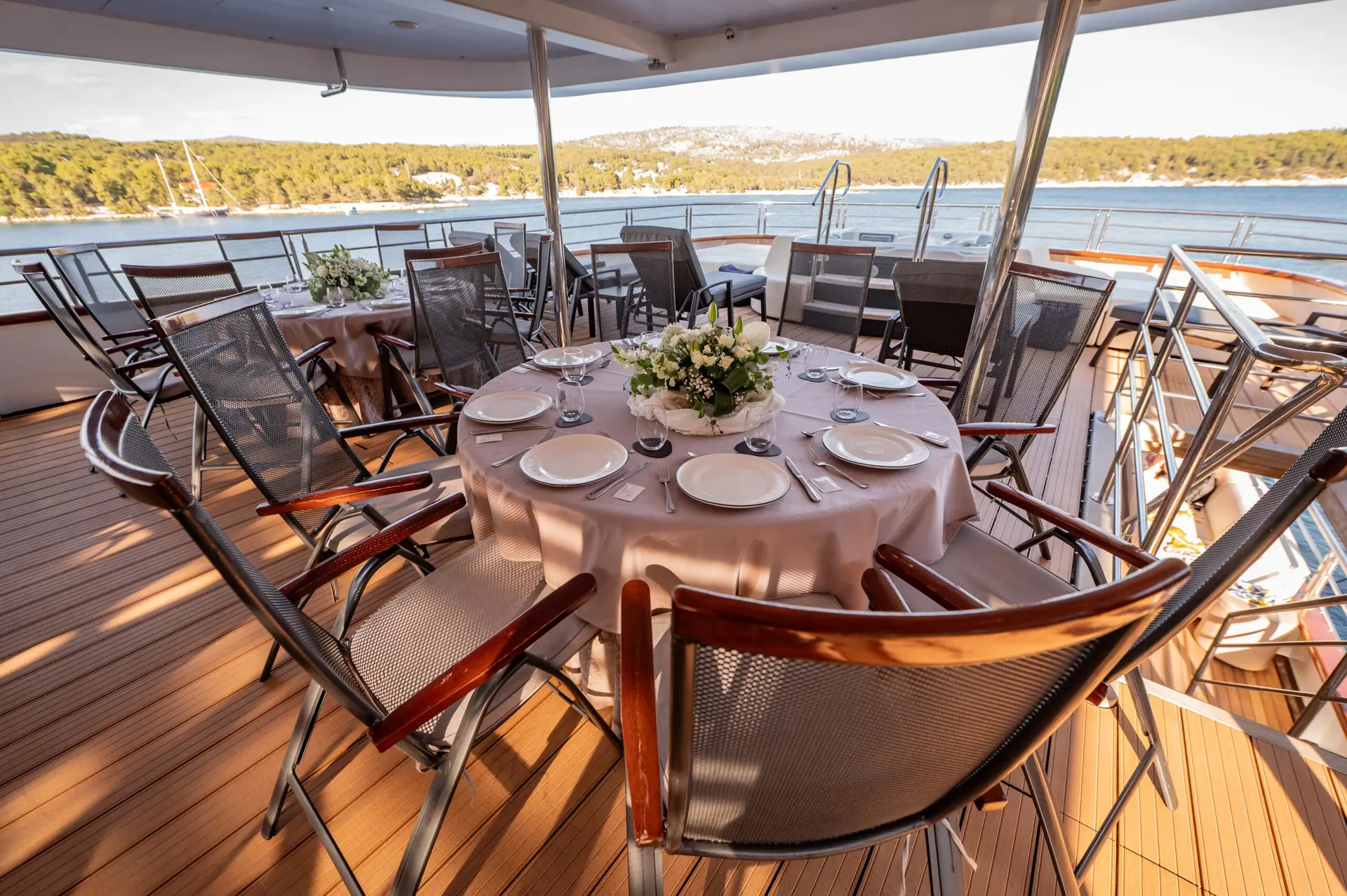 A round table set for dining with eight chairs on a yachts deck, overlooking calm water and a wooded shoreline in the background under a clear sky.