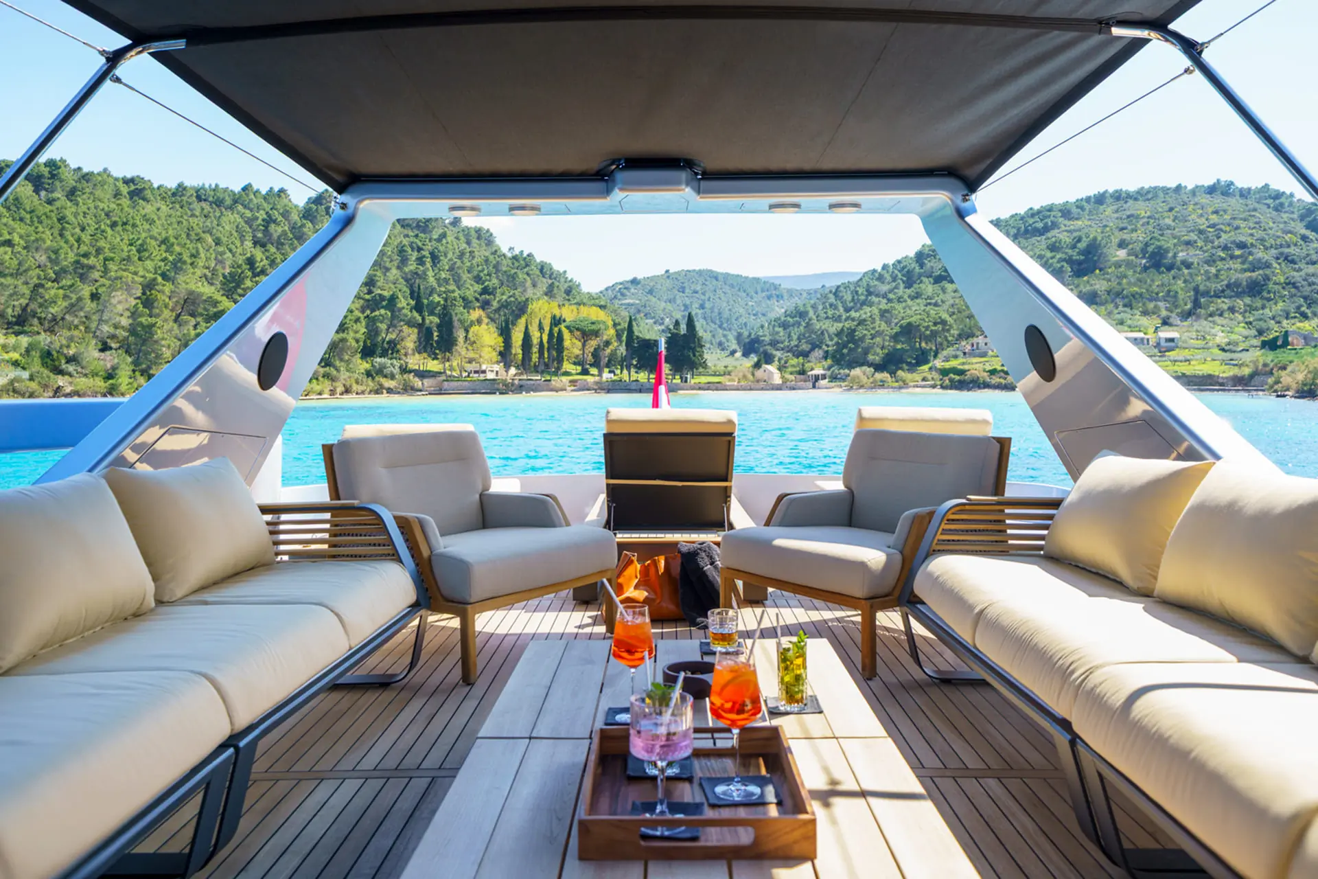 A spacious yacht deck with beige seating, a table with drinks, and a view of blue water and green hills in the background under a sunny sky.