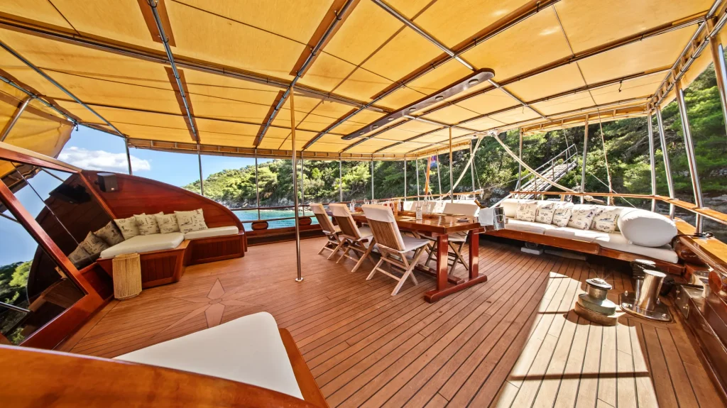 ELEGANZA A spacious yacht deck with wooden flooring, cushioned seating, a dining table with chairs, and a yellow canopy overhead. The deck overlooks clear blue water and a lush green shoreline in the background.