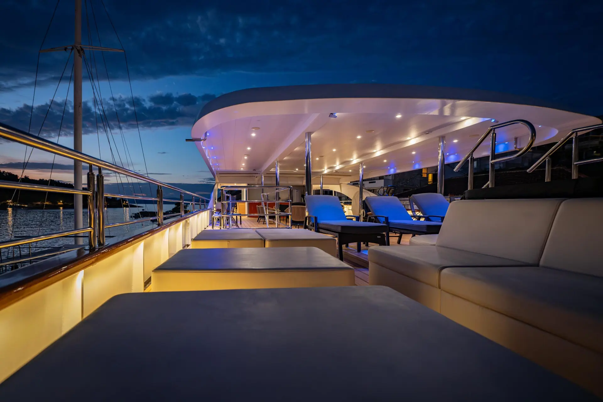 Luxury yacht deck at dusk with modern lounge chairs, white sofas, warm lighting, and a calm ocean in the background under a twilight sky.