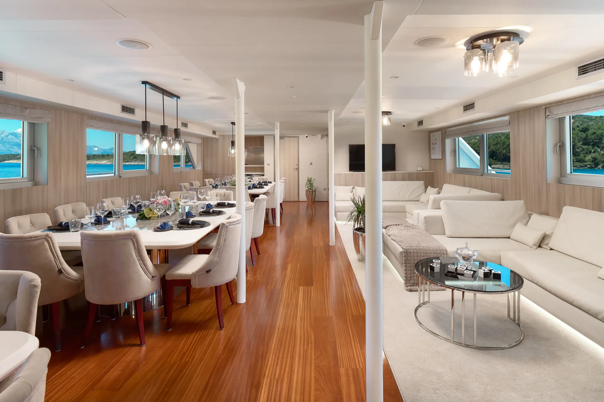 CRISTAL A spacious, modern yacht interior with wooden floors, white walls, and large windows. It features elegant dining tables with set places, plush chairs, and a cozy lounge area with white sofas and a coffee table.