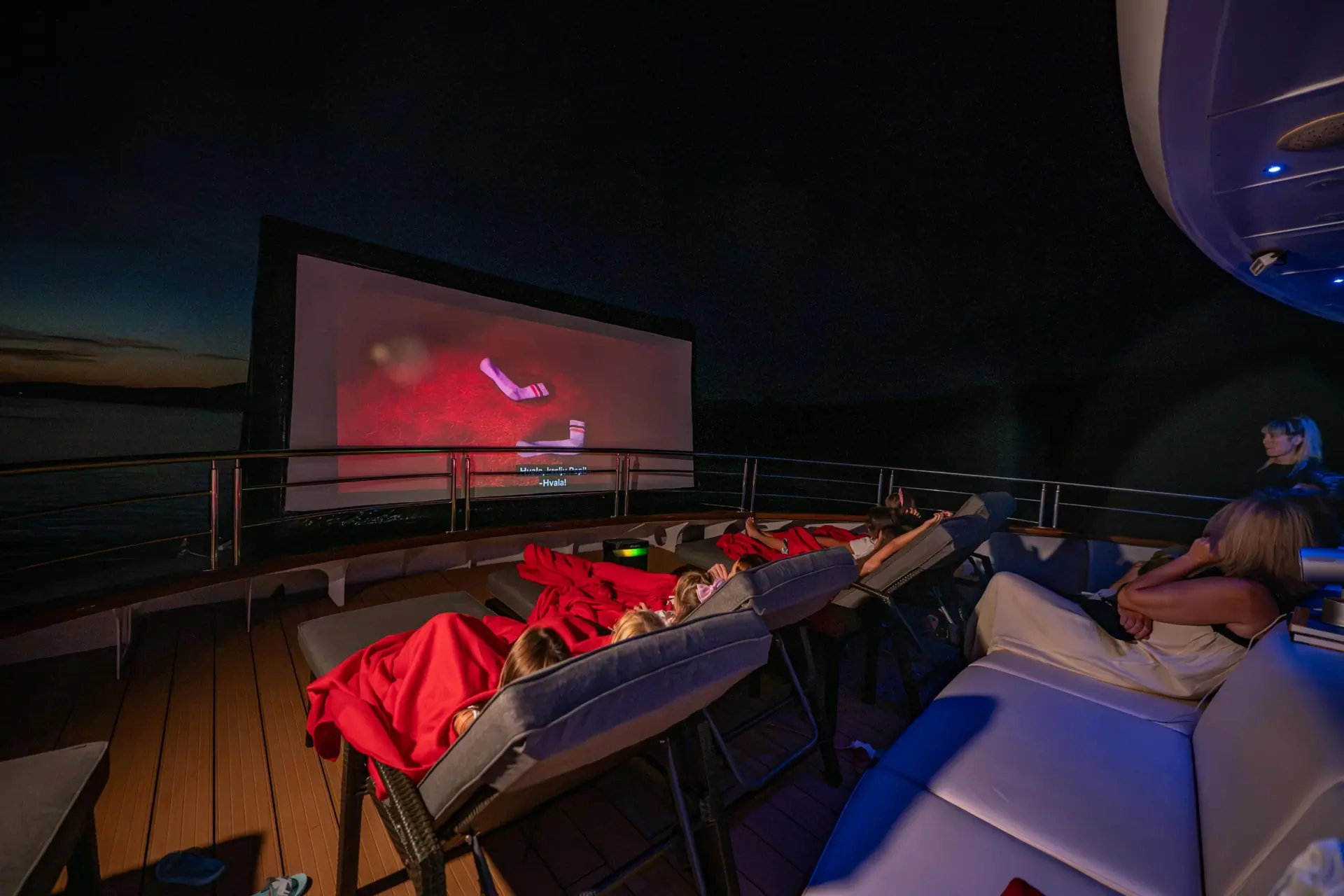 Several people lounge on deck chairs under red blankets, watching a movie on a large outdoor screen at night on a yacht. The scene is dimly lit, creating a cozy, relaxed atmosphere.
