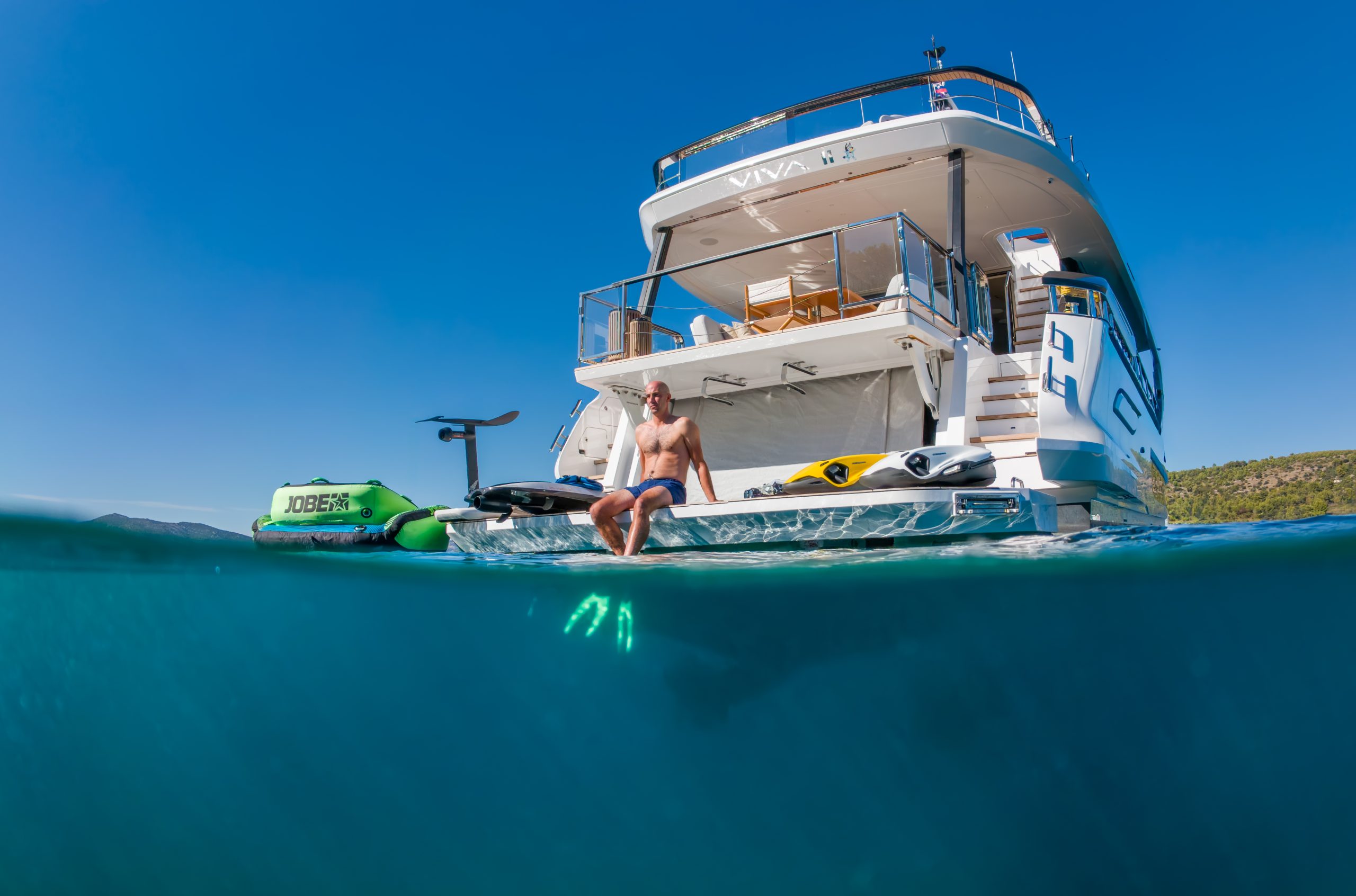VIVA II A man in swim trunks sits on the back deck of a yacht, with water toys nearby. The photo is half underwater, showing clear blue water below and the boat above, under a bright blue sky.