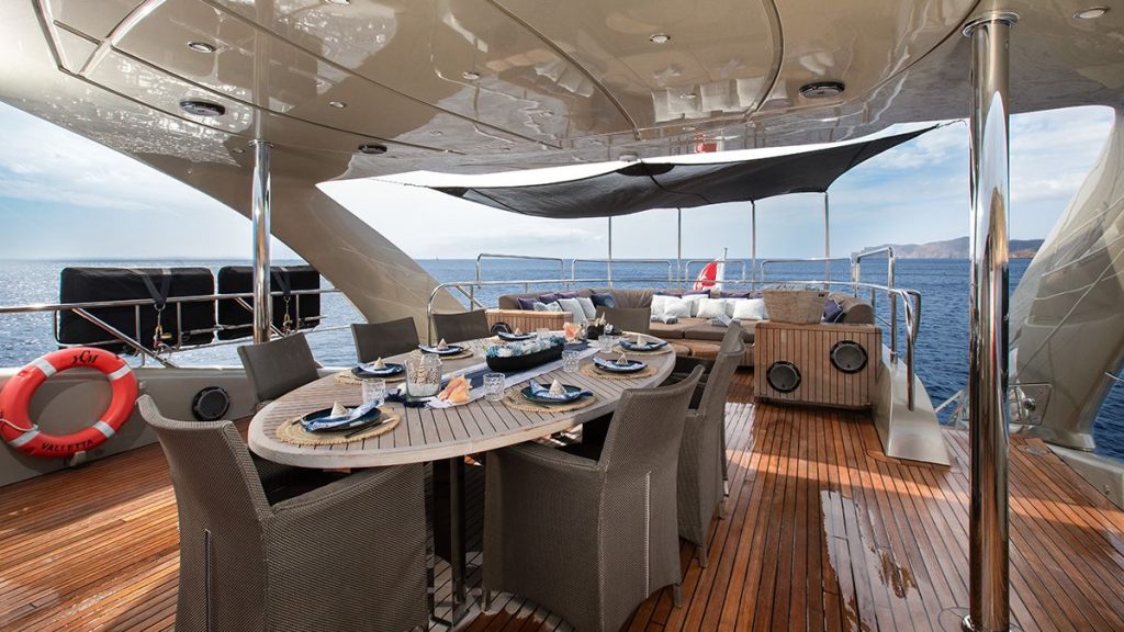 YCM 120 Elegant yacht deck with a wooden dining table set for eight, cushioned seating area in the background, and expansive ocean views under a partly shaded canopy.