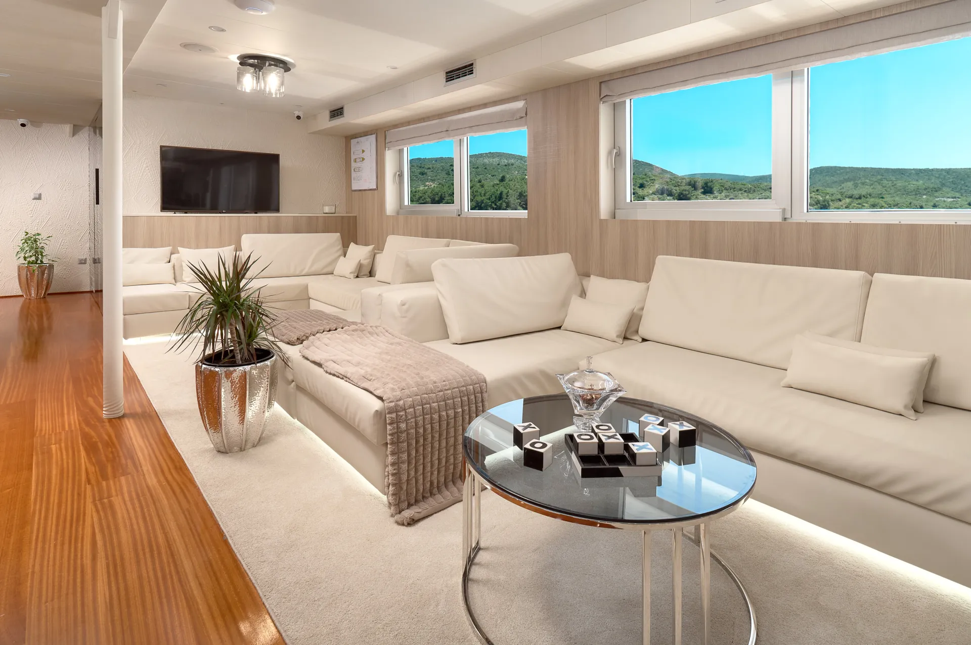 CRISTAL A modern living room with white sofas, a glass coffee table, and a plush beige throw blanket. Large windows offer a view of green hills and blue sky, and a TV is mounted on the wall. A potted plant and decor items adorn the space.