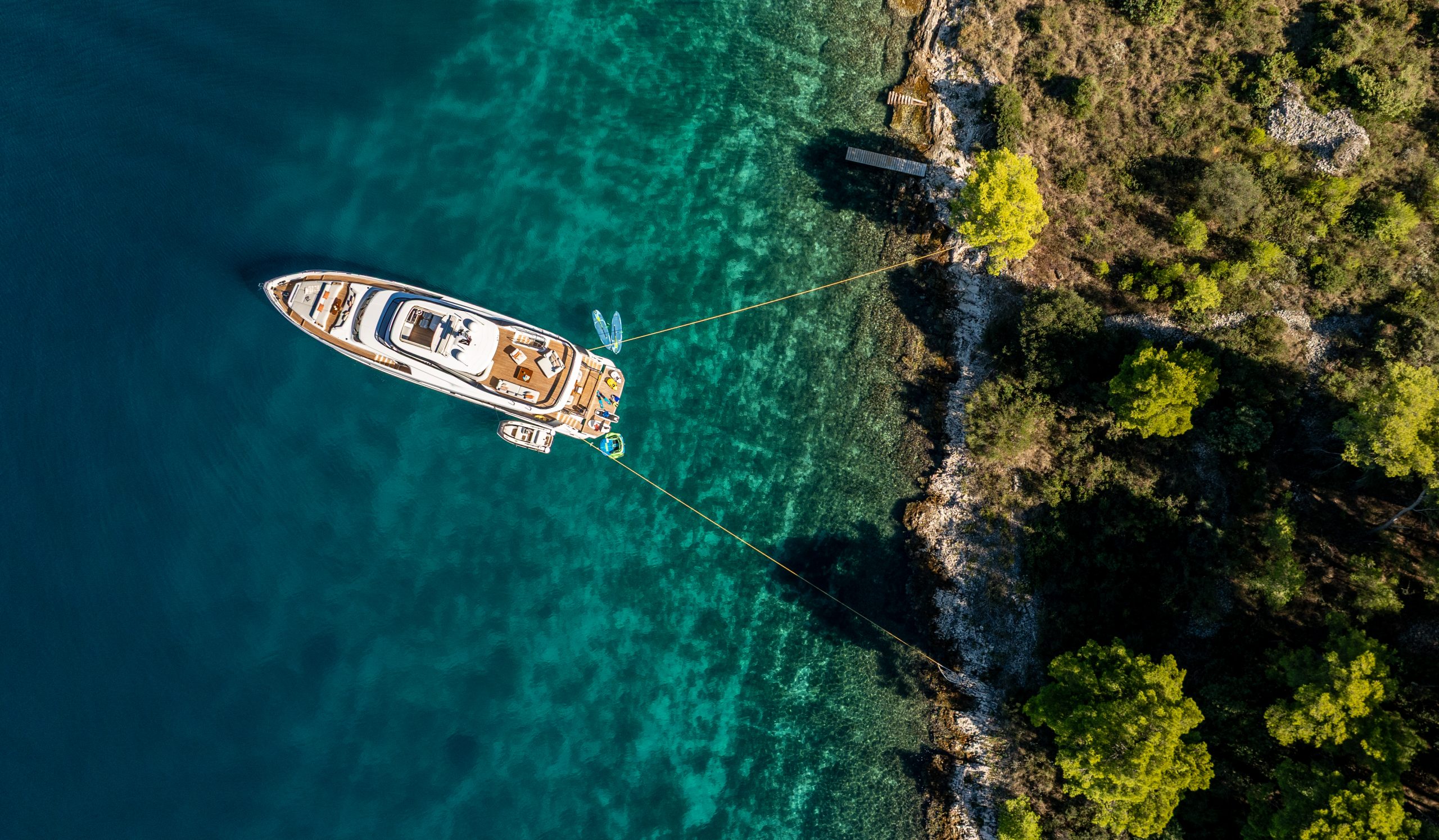 VIVA II Aerial view of a luxury yacht anchored in clear turquoise water, tethered to a rocky, tree-lined shoreline with lush green vegetation.