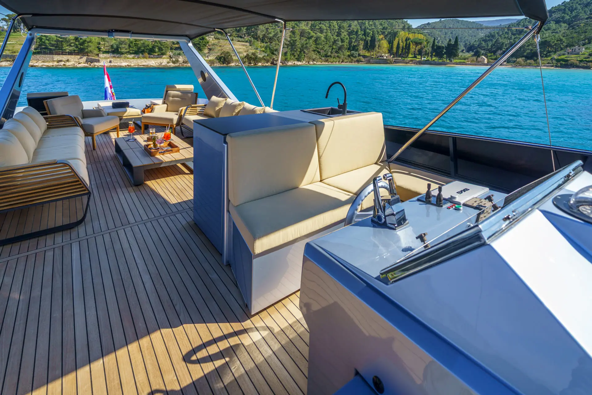 The upper deck of a luxury yacht with cushioned seating, wooden flooring, a control panel, and views of blue water and green trees in the background.