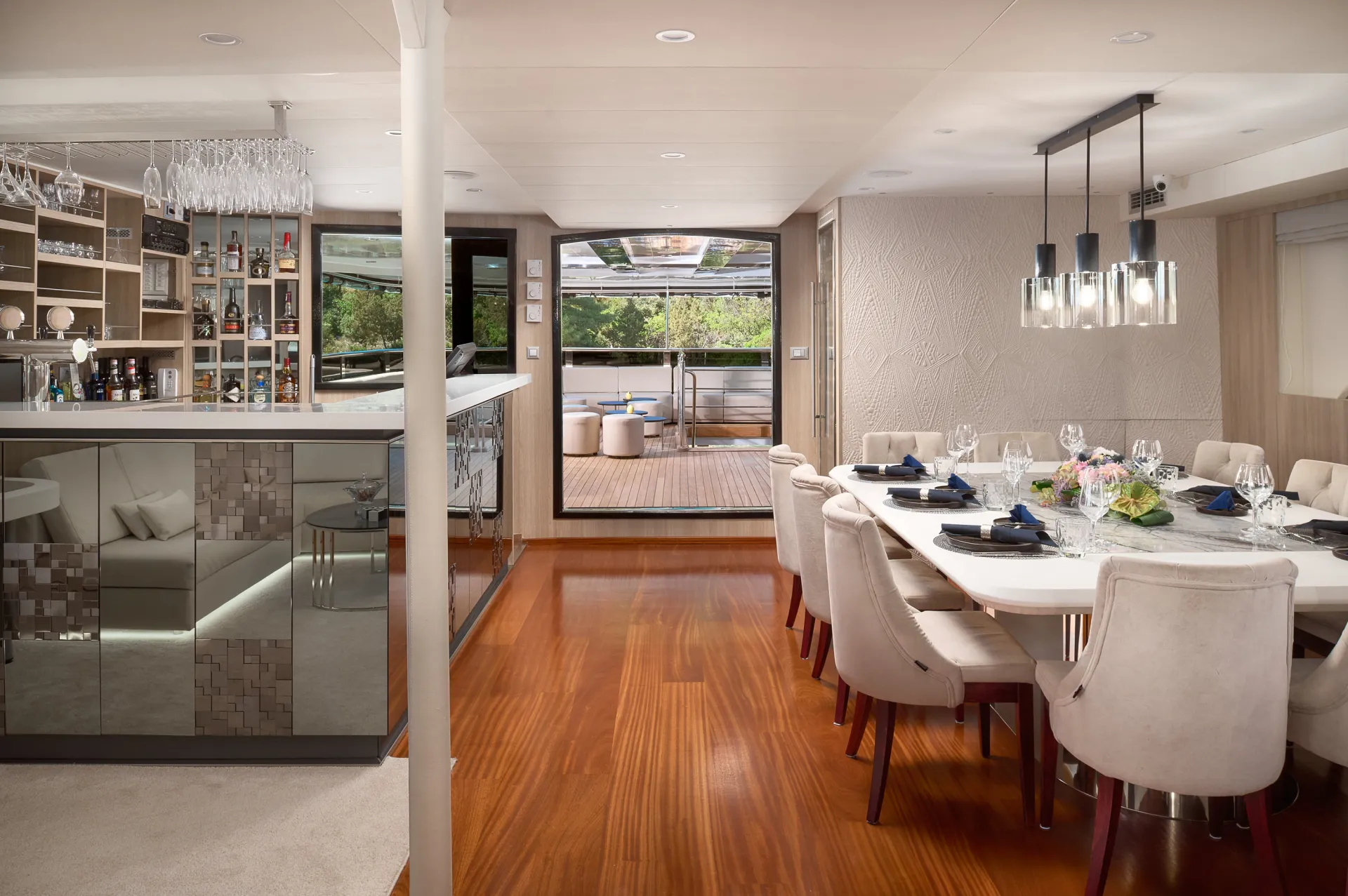 CRISTAL Modern dining area on a yacht with a long white table set for a meal, plush beige chairs, polished wood floor, mirrored bar with glassware, and an open door leading to an outdoor deck with seating.