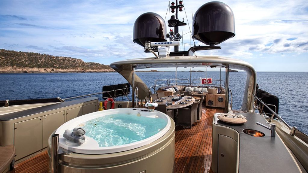 YCM 120 A luxury yacht deck with a round hot tub, outdoor dining table set for a meal, lounge seating, and a scenic ocean view with distant hills under a partly cloudy sky.