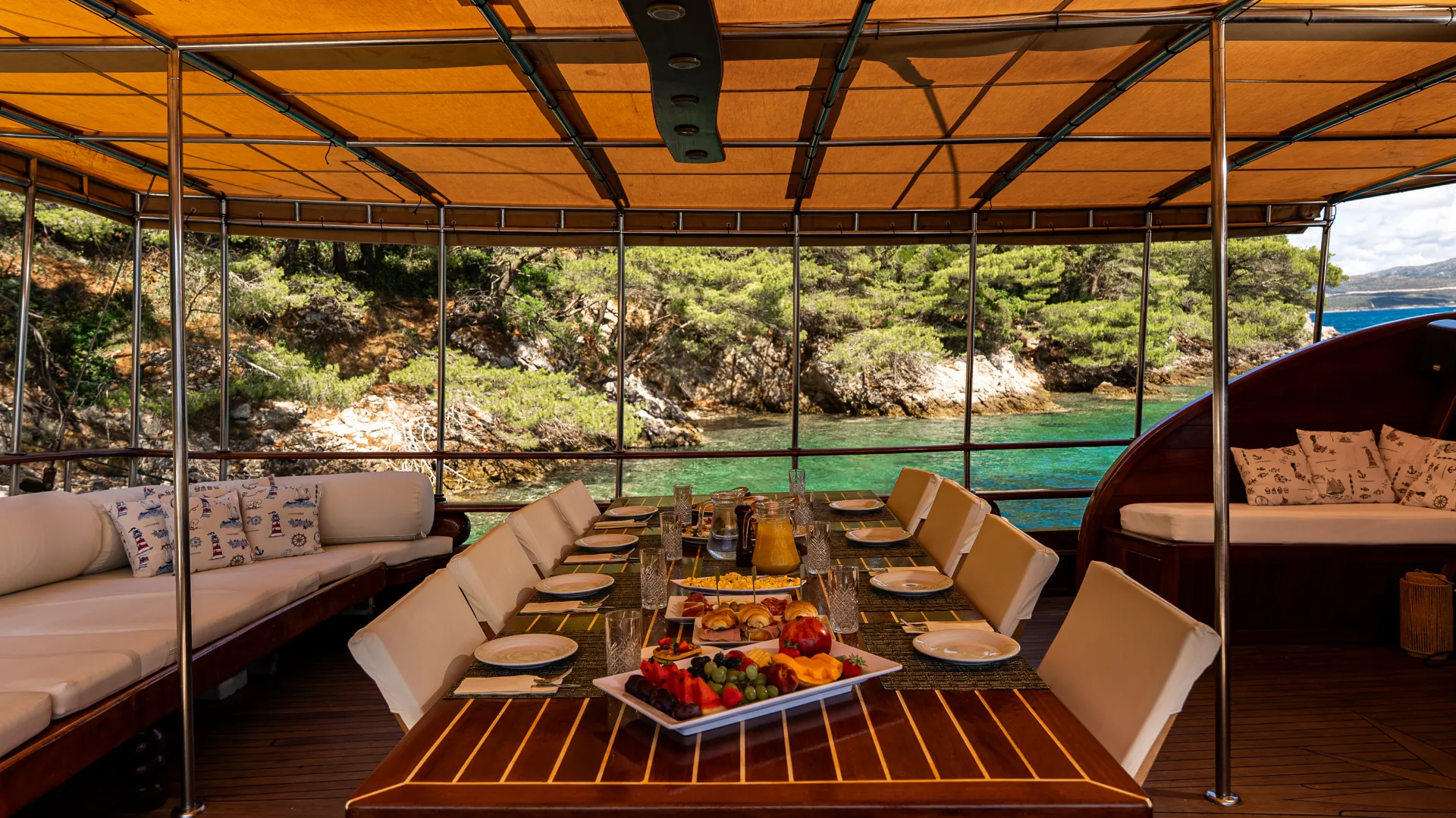 ANDI STAR An outdoor dining area on a yacht with a wooden table set for a meal, featuring plates, glasses, and fruit platters. The yacht is anchored near clear turquoise water and a rocky, forested shoreline.