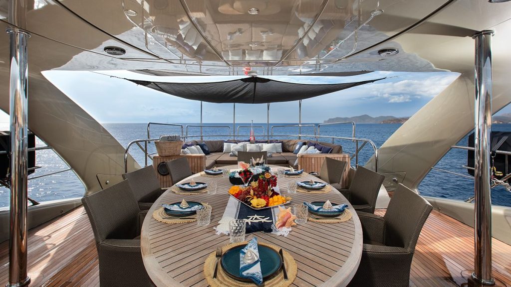 YCM 120 A luxurious yacht deck with an elegant dining table set for eight, decorated with flowers and napkins, overlooking calm blue sea and distant hills under a partly cloudy sky.