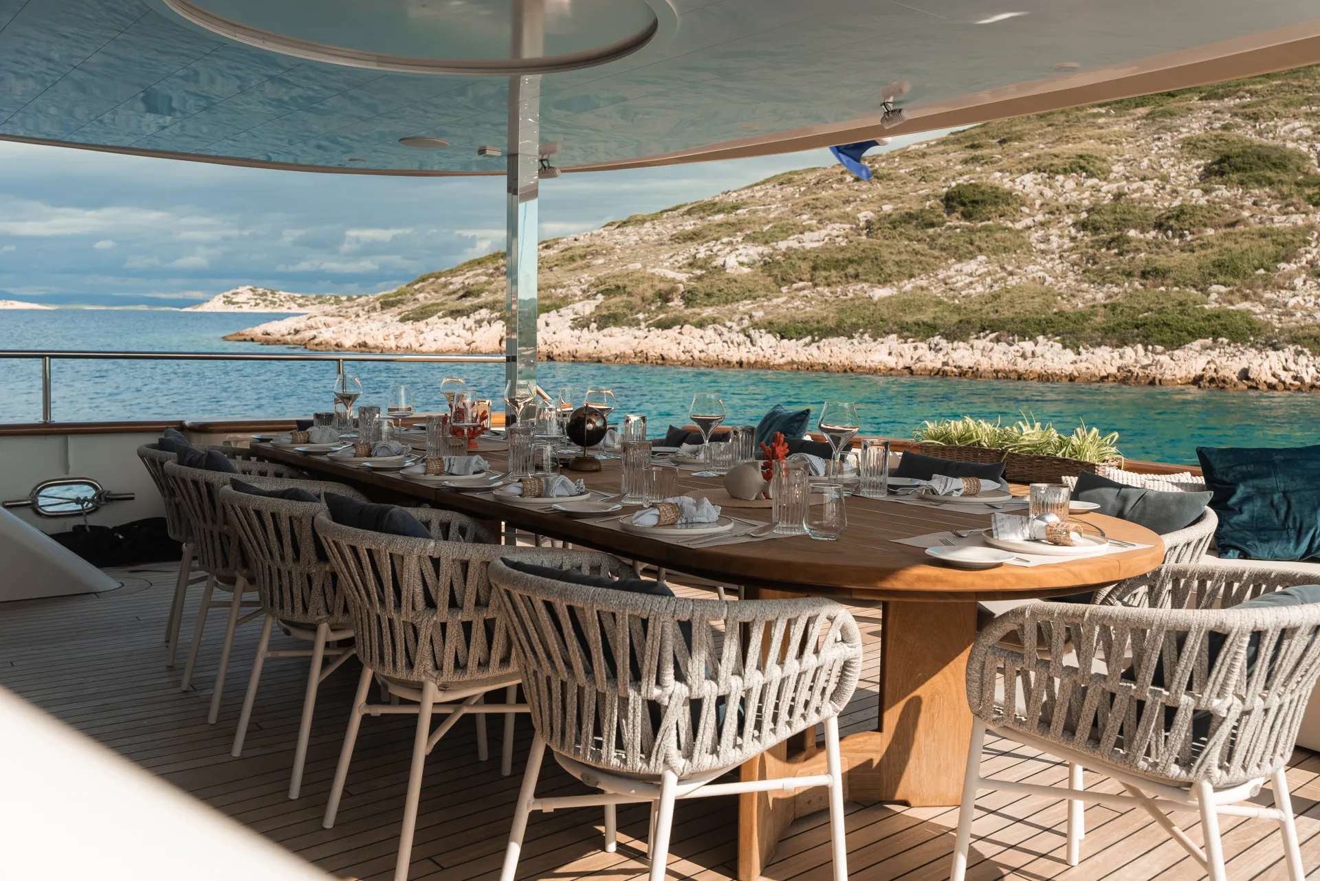 MAXITA A large outdoor dining table set for a meal sits on the deck of a yacht, surrounded by cushioned chairs, overlooking turquoise water and a rocky, green shoreline under a partly cloudy sky.