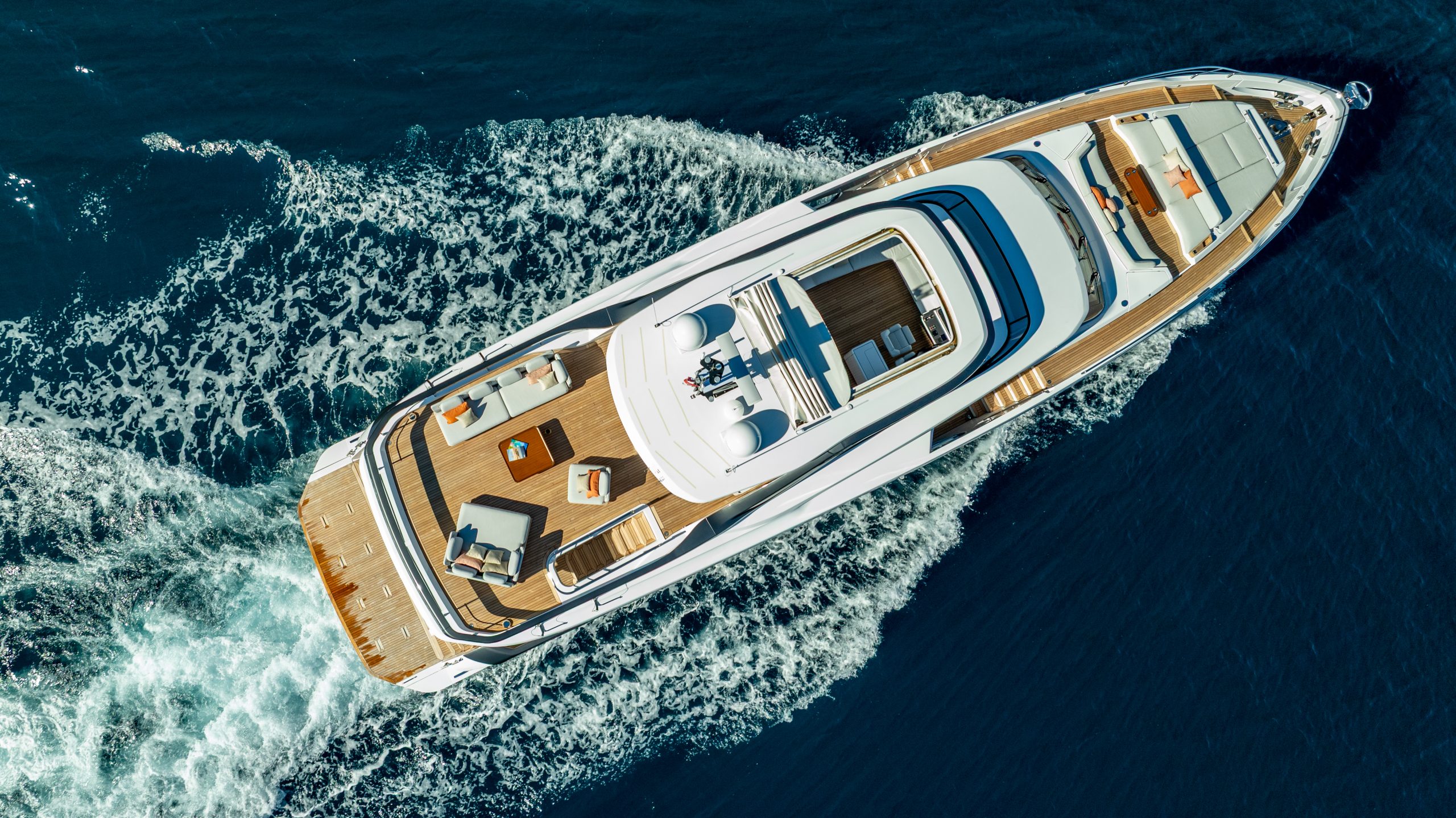 VIVA II Aerial view of a luxury yacht cruising through deep blue water, leaving a white foamy wake behind. The yacht’s deck features seating areas, sunbeds, and lounging spaces.