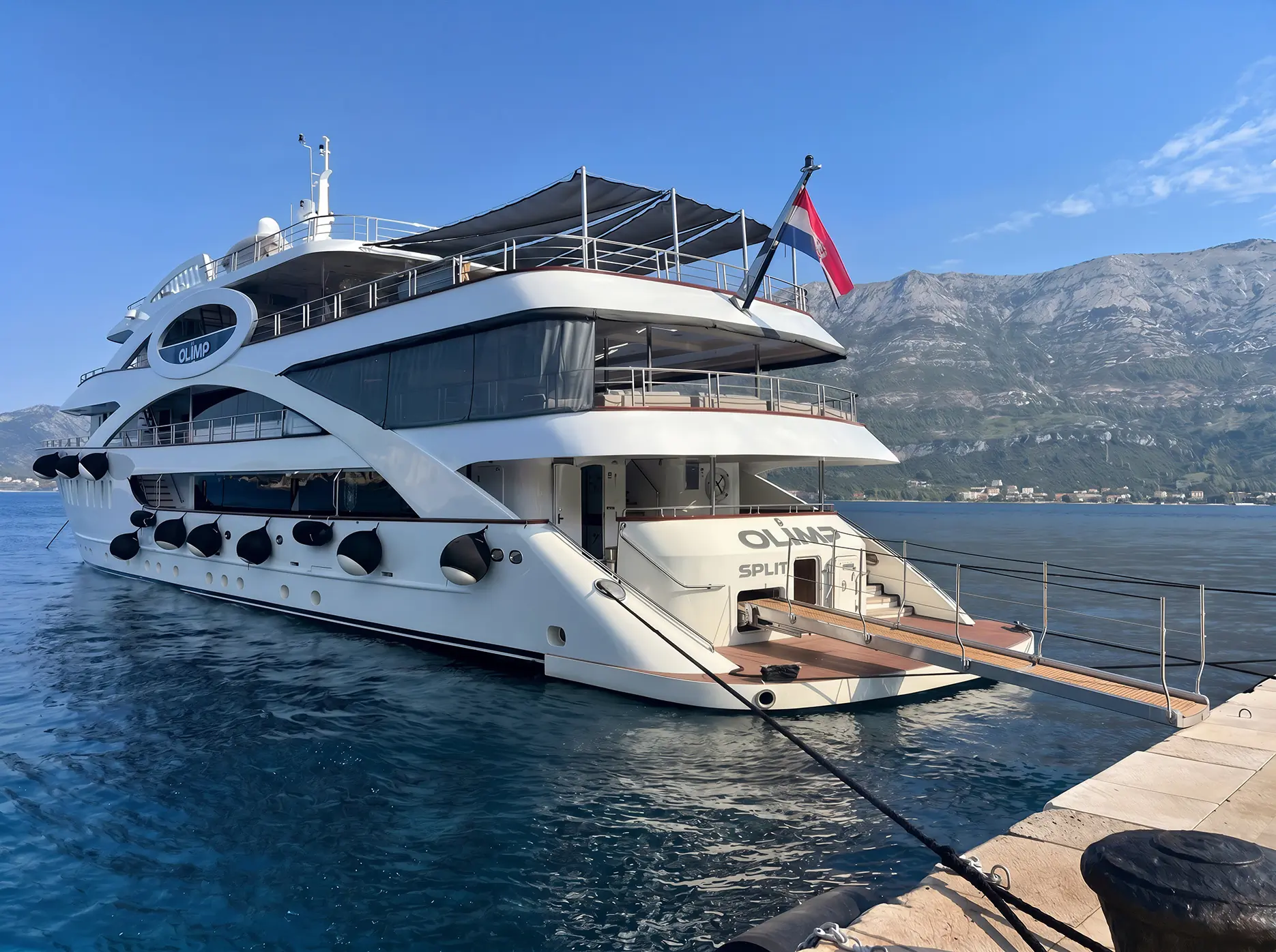 A large white yacht named Olimp is docked at a pier, with mountains and blue sky in the background. The yacht has multiple decks and a red-and-white flag flying at the stern.