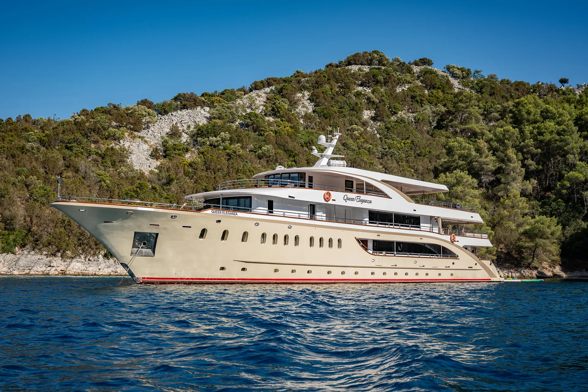 A large luxury yacht with multiple decks floats on calm blue water, with a green, forested hillside in the background under a clear blue sky.