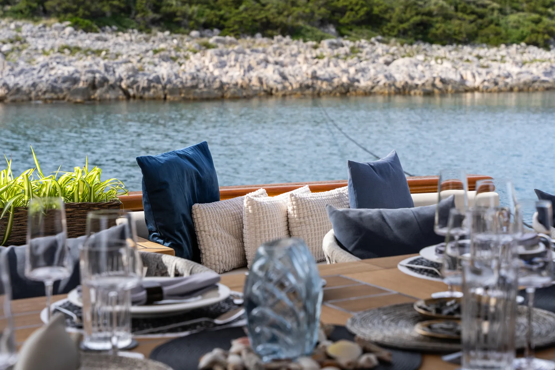 MAXITA A waterfront outdoor dining table set with glassware, plates, and decorative items, with cushioned seating and pillows in the background overlooking calm water and a rocky shoreline.