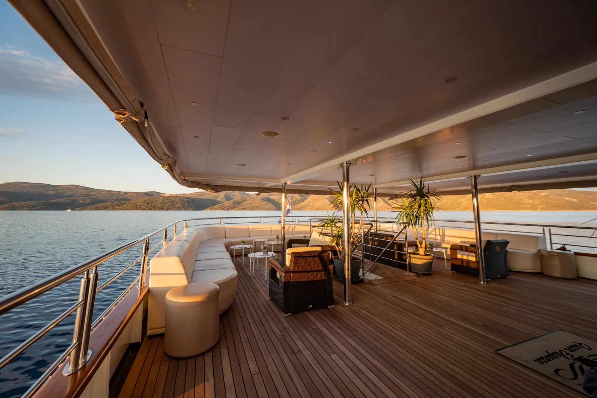 Spacious yacht deck with wooden flooring, white cushioned seating, potted plants, and outdoor furniture, overlooking calm water and distant hills at sunset.