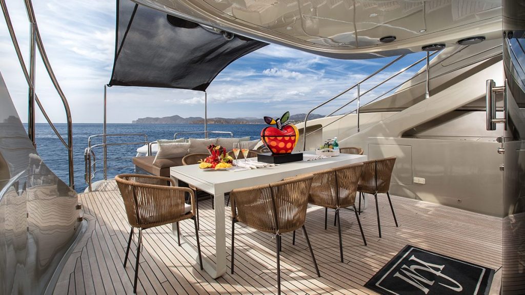 YCM 120 A modern yacht deck with a dining table set for four, wicker chairs, fruit centerpiece, and ocean view under a partly shaded canopy. Distant islands and a blue sky are visible in the background.