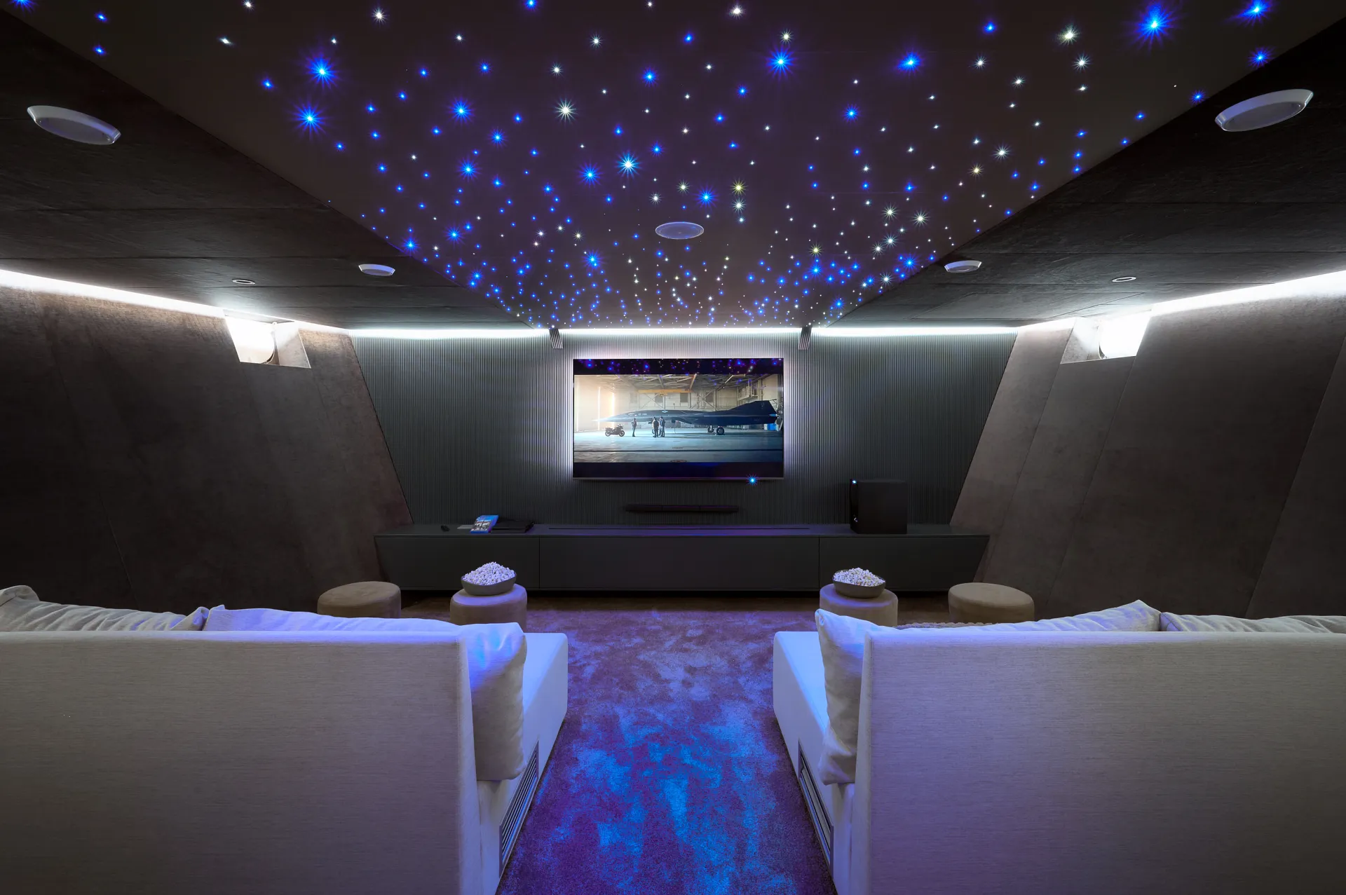 CRISTAL A modern home theater room with two white sofas facing a large screen, star-like LED lights on the ceiling, dark walls, and soft, plush carpet flooring.