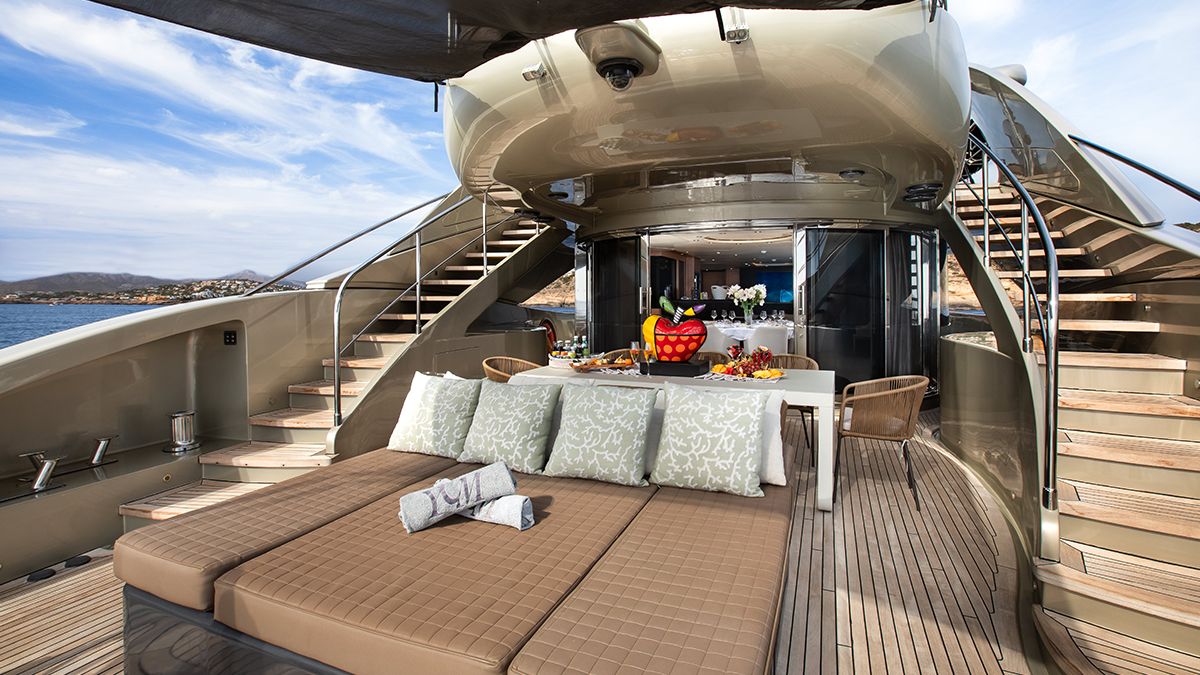 YCM 120 Luxurious yacht deck with cushioned sunbeds, a table set with fruit and drinks, and outdoor seating; stairs lead up on both sides, and the sea is visible in the background under a partly cloudy sky.
