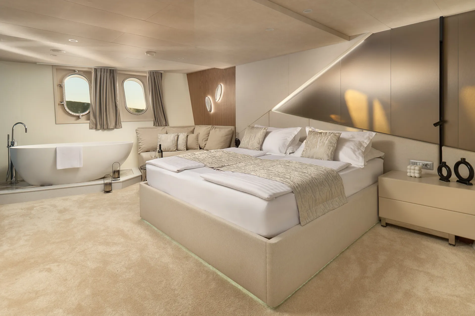 CRISTAL A spacious, modern bedroom with a large bed, beige carpet, a freestanding bathtub near two round windows, a cozy corner sofa, and minimalist decor in neutral tones.