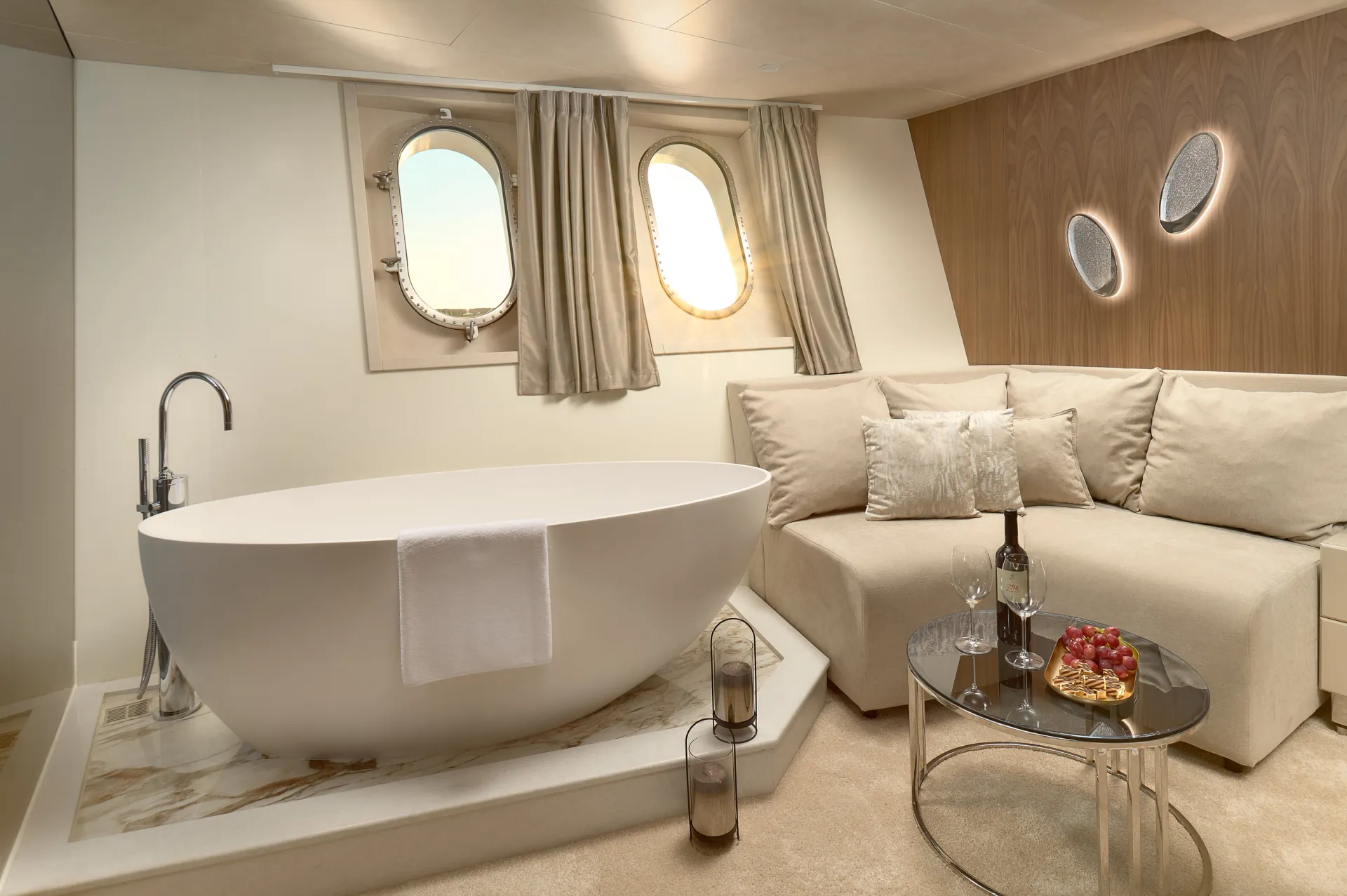 CRISTAL A luxurious bathroom with a modern white bathtub, a towel draped over the side, porthole windows, a beige couch with pillows, a round glass table holding wine, glasses, and grapes, and soft, neutral decor.