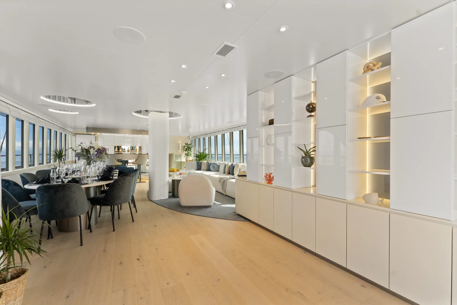 MAXITA Luxurious yacht interior with modern white decor, open shelving, and elegant dining area. Large windows provide ample natural light. There are plants and decorative items on display, with a cozy seating area in the background.